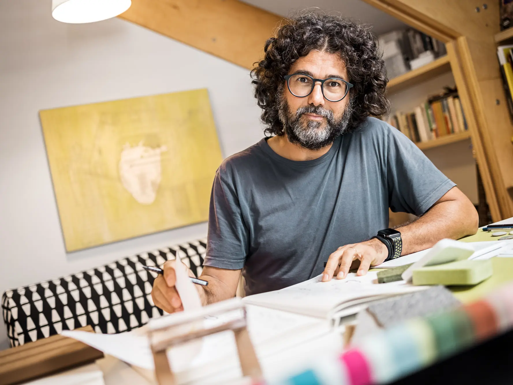 Portrait de l’architecte Sebastiano Longaretti chez lui, dans son atelier, avec un de ses tableaux accroché au mur derrière lui. Stylo à la main, il feuillette un carnet de dessin.