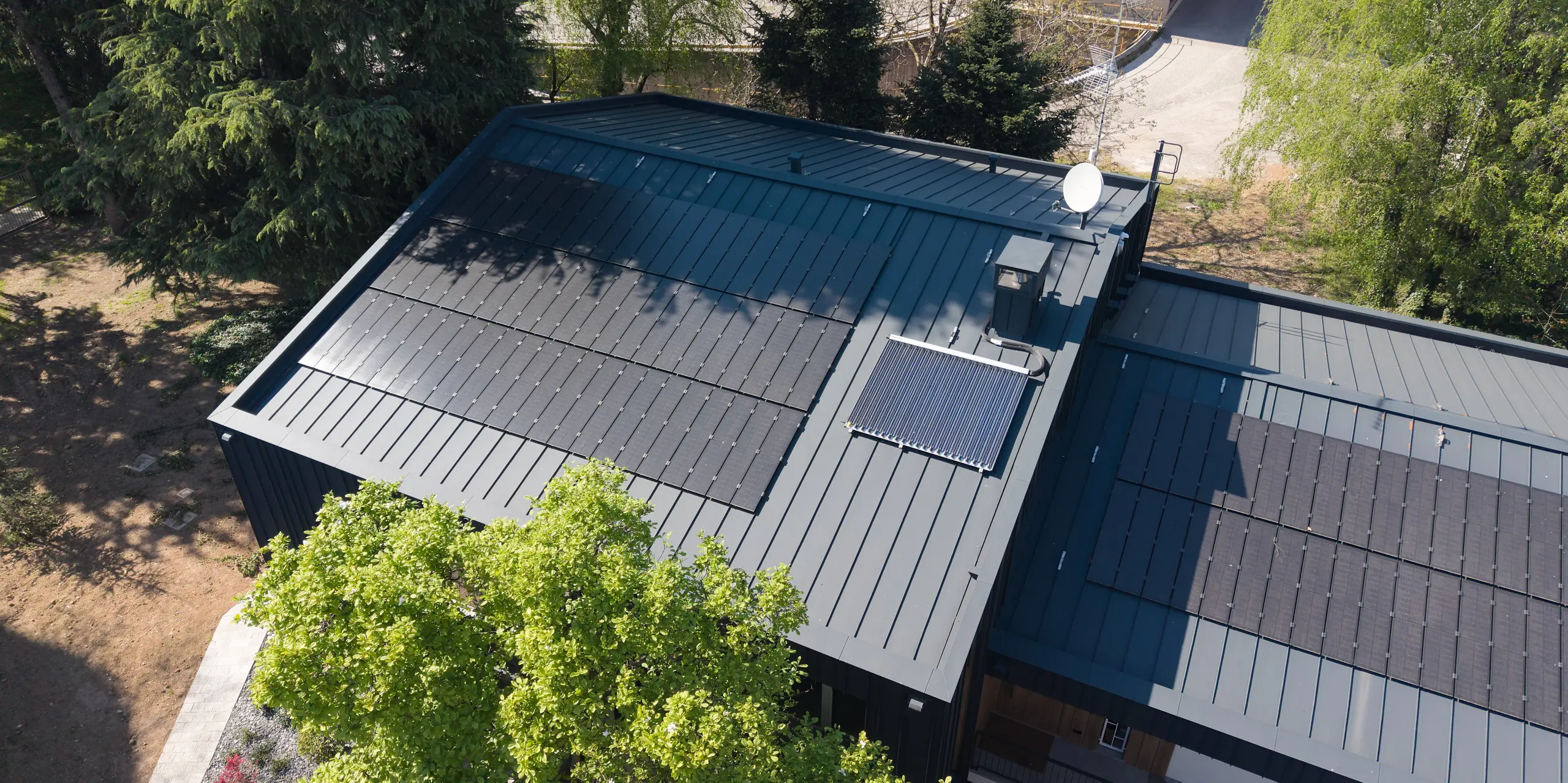 Vue aérienne de la Villa Castellanza, dotée d'une toiture PREFALZ couleur Anthracite P.10 et de modules solaires PREFALZ parfaitement intégrés. La pureté des joints debout souligne l'architecture moderne et met en valeur la qualité de l'enveloppe du bâtiment PREFA. Entourée d'arbres matures, la propriété allie avec élégance production d'énergie durable, robustesse de l'aluminium et esthétique intemporelle.