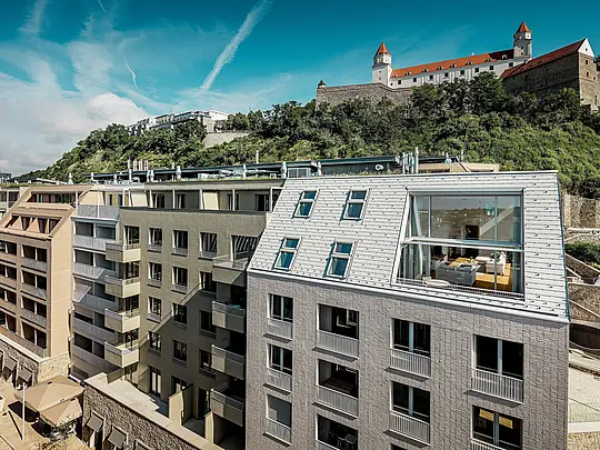 Une toiture moderne dans le quartier de Vydrica à Bratislava, offrant une vue imprenable sur le château : les toitures à forte pente, percées de larges ouvertures vitrées et aux lignes épurées, sont élégamment mises en valeur par les tuiles en aluminium PREFA, disponibles en blanc Prefa et brun sable. La précision de l’exécution et l’intégration harmonieuse de la toiture et de la façade créent un point focal visuel saisissant au-dessus des toits de la ville. L’aluminium PREFA séduit par sa légèreté, sa grande résistance aux intempéries et sa durabilité – un matériau idéal pour les immeubles résidentiels complexes et à l’architecture raffinée.