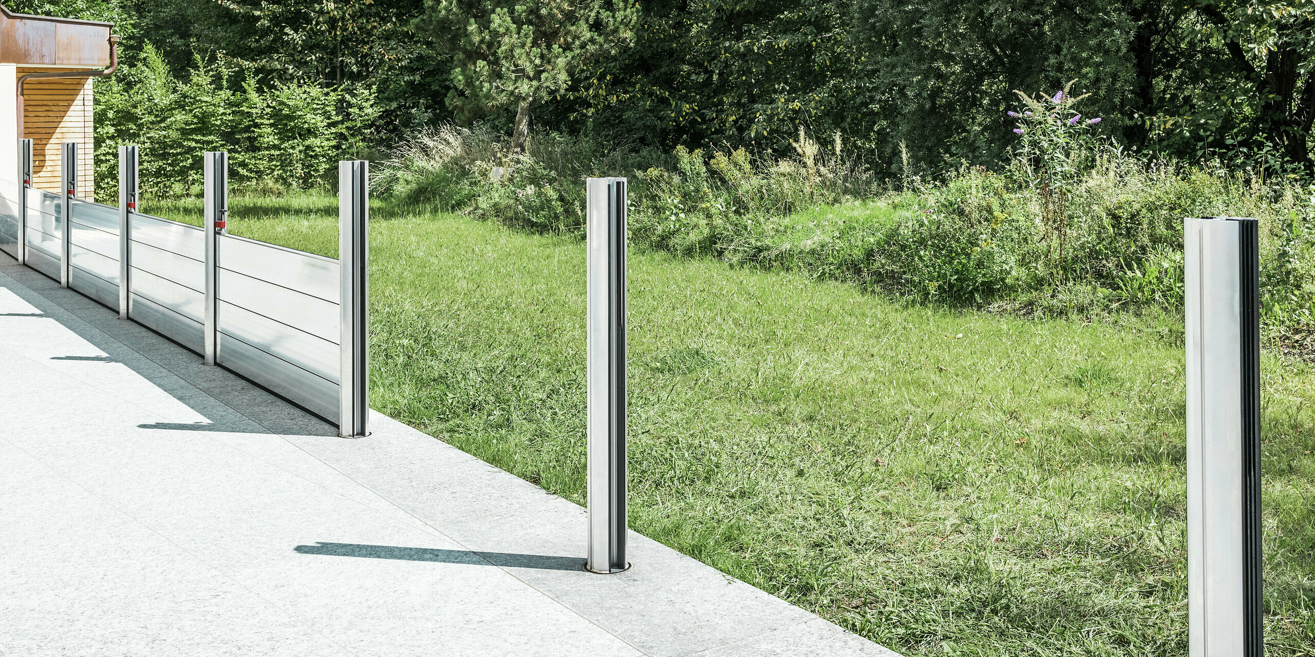 Vue détaillée du système mobile de protection contre les crues PREFA en cours d'installation sur une terrasse au Tyrol. Les douilles de sol déjà installés et les supports en aluminium insérés qui soutiennent le système de batardeaux sont visibles. En arrière-plan, on distingue la prairie adjacente et le terrain dégagé – une application typique de la protection contre les crues PREFA. La construction en aluminium de haute qualité offre une protection fiable contre les inondations et une esthétique élégante. Aucun batardeau n'a encore été inséré dans les deux supports avant, le système étant encore en phase d'installation au moment de la photo.