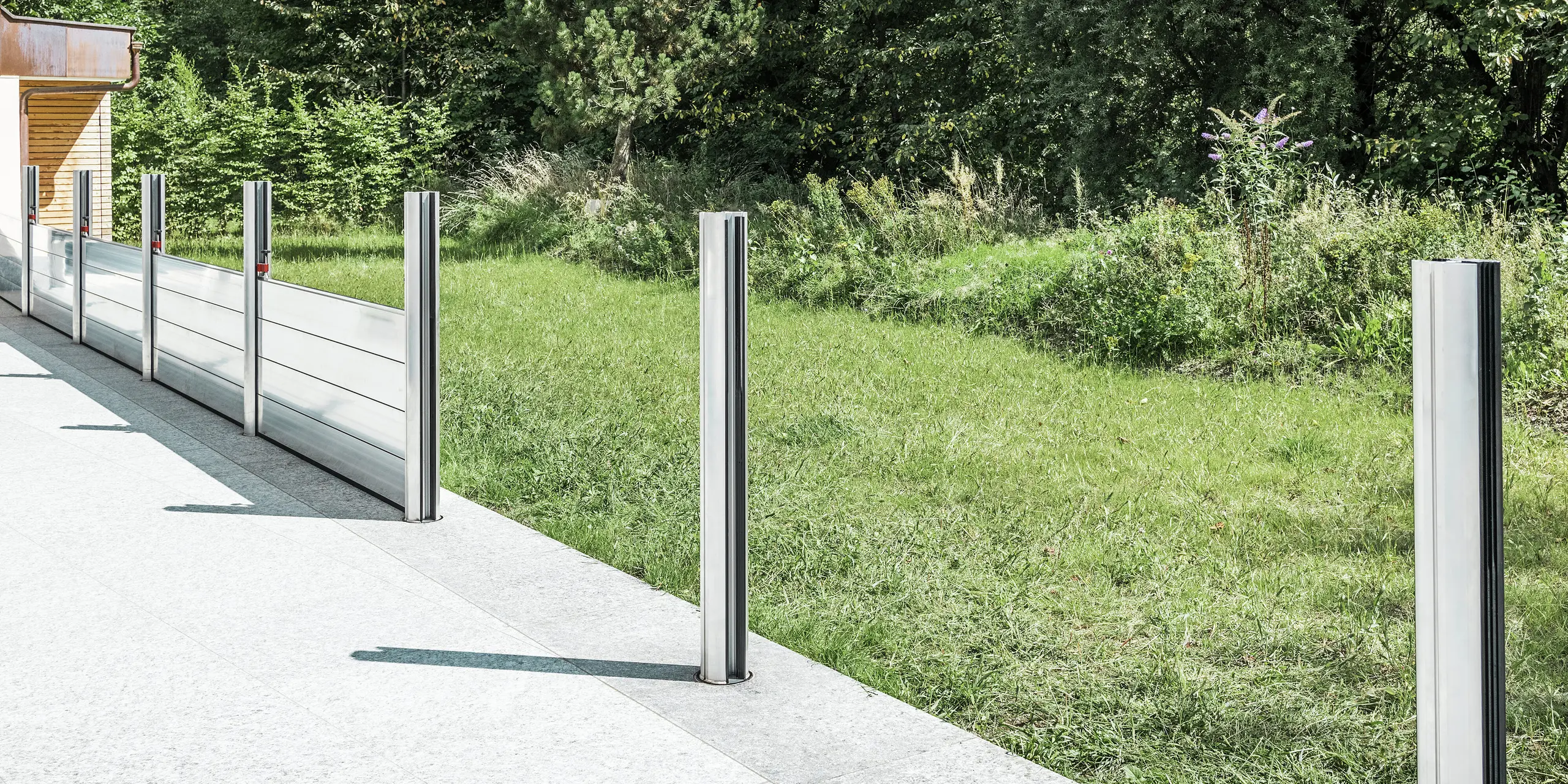 Vue détaillée du système mobile de protection contre les crues PREFA en cours d'installation sur une terrasse au Tyrol. Les douilles de sol déjà installés et les supports en aluminium insérés qui soutiennent le système de batardeaux sont visibles. En arrière-plan, on distingue la prairie adjacente et le terrain dégagé – une application typique de la protection contre les crues PREFA. La construction en aluminium de haute qualité offre une protection fiable contre les inondations et une esthétique élégante. Aucun batardeau n'a encore été inséré dans les deux supports avant, le système étant encore en phase d'installation au moment de la photo.