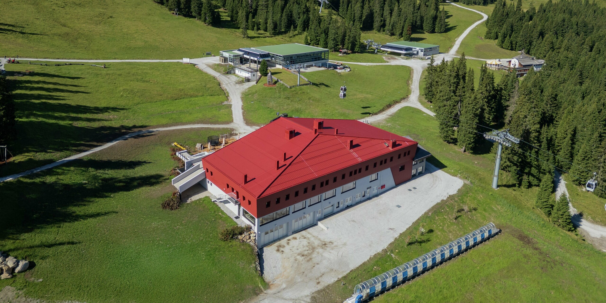 L'imposant bâtiment du Summit Mountain Club à Ratschings se détache avec force sur le paysage alpin. Son revêtement en aluminium rouge oxyde, avec la marque PREFALZ sur la toiture et les bardeaux PREFA sur la façade, attire particulièrement le regard. Au premier plan, on aperçoit de vastes prairies et une route d'accès au bâtiment. La station de remontées mécaniques et le vaste domaine à l'arrière-plan soulignent le panorama montagneux, tandis que le bâtiment lui-même, avec son design moderne et fonctionnel, abrite à la fois un restaurant et un espace événementiel.
