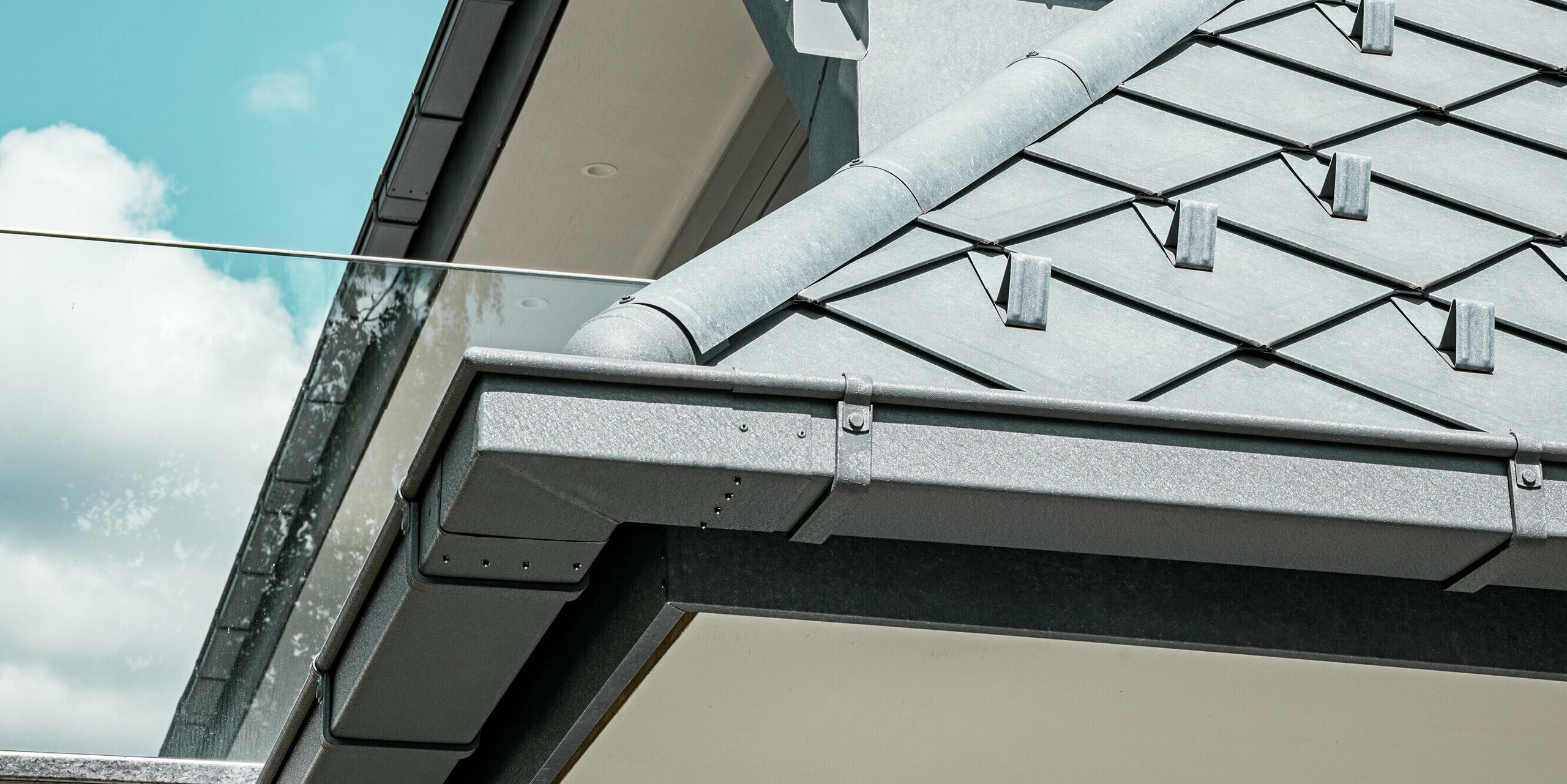 Gros plan sur la toiture d'un immeuble d'appartements moderne, revêtue de losanges PREFA 29 x 29 couleur Gris Pierre P.10. La rive de toit est élégamment finie avec une gouttière profilée et une descente d'eau pluviale en aluminium, assurant une évacuation efficace des eaux. Le balcon vitré complète le design minimaliste du bâtiment et contraste avec la robustesse de la charpente. La qualité des matériaux et la précision de la réalisation offrent des avantages à la fois esthétiques et fonctionnels, contribuant à un cadre de vie moderne et agréable.