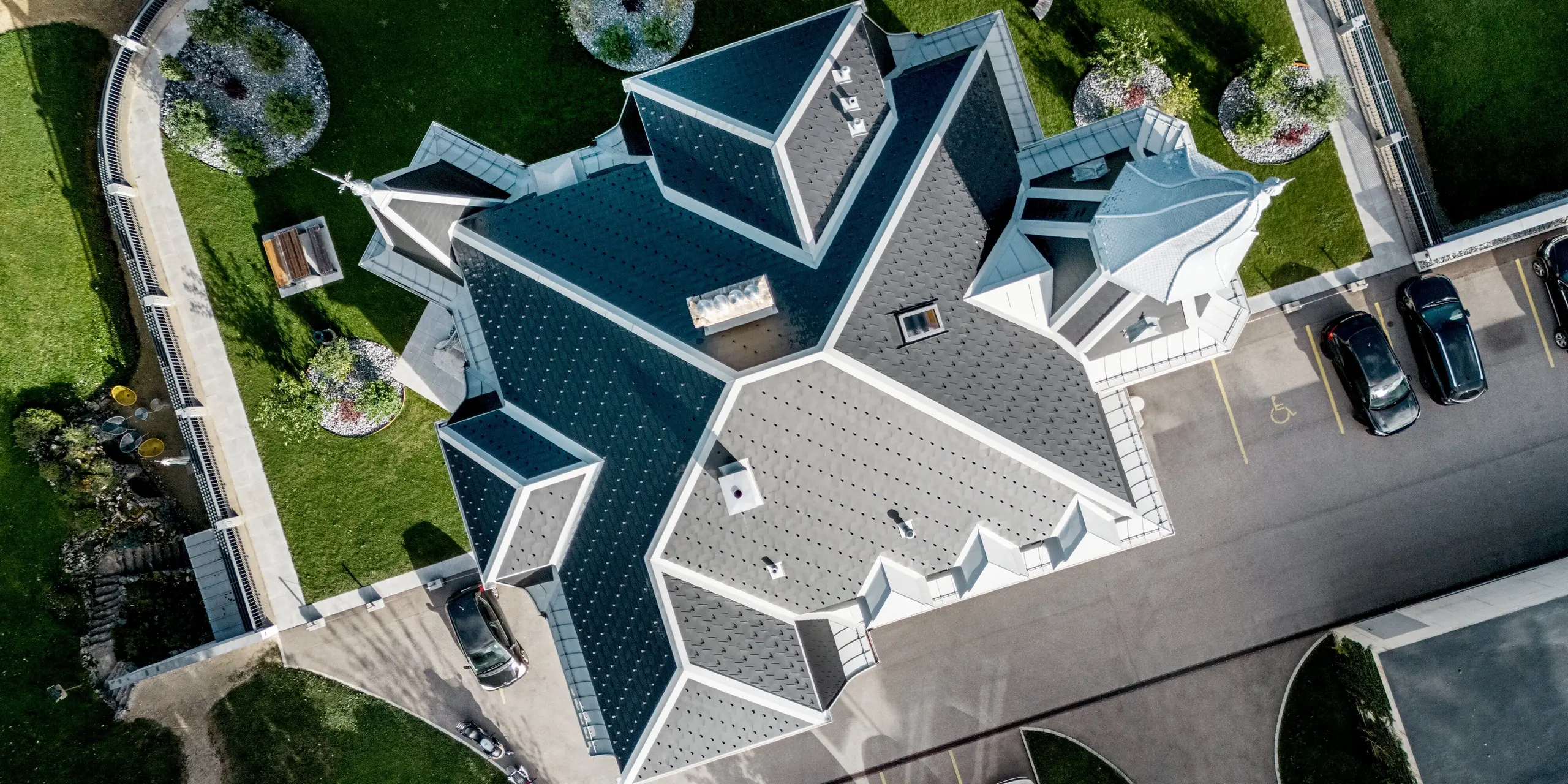 Dramatische Drohnenaufnahme der Manufacture Horlogère du Locle in der Schweiz mit spektakulärer, vielgliedriger Dachlandschaft. Die komplexen Dachflächen, markanten Türme und geschwungenen Übergänge wirken wie eine architektonische Skulptur und werden durch PREFA Dachschindeln in P.10 Schwarz eindrucksvoll betont. PREFALZ in Patinagrau sorgt für präzise Ausführung bei Rundungen und Anschlüssen. Das leichte Aluminiumdach überzeugt durch Langlebigkeit, Widerstandsfähigkeit und flexible Gestaltung, während umliegende Grünflächen und Wege die außergewöhnliche Form zusätzlich in Szene setzen.