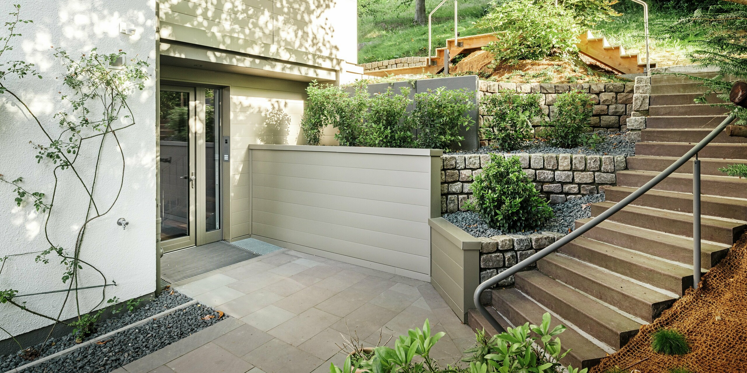 L'entrée de la Villa Blankenese à Hambourg illustre parfaitement l'harmonie entre architecture moderne et matériaux haut de gamme. La pureté des lignes et l'alliance de la pierre naturelle, de la verdure et du métal créent une impression d'élégance. Les éléments de façade couleur bronze, réalisés en Siding PREFA, sont particulièrement remarquables et confèrent au bâtiment un caractère à la fois chaleureux et raffiné.