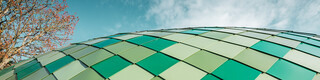 Gros plan sur la façade en aluminium à courbures multiples dans trois tons de vert différents de PREFA, le ciel et un arbre sont visibles en arrière-plan.