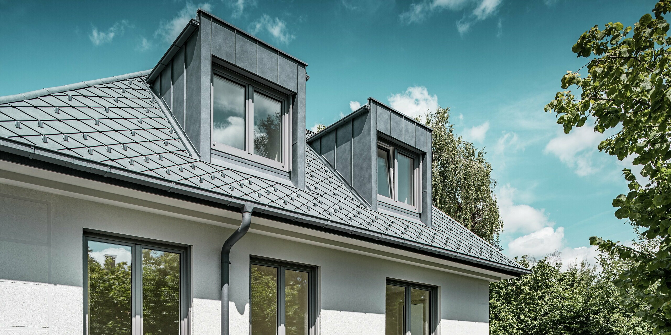 Une élégante perspective d'un immeuble d'appartements aux remarquables lucarnes à toit plat en PREFALZ et à la toiture parfaitement agencée, composée de losanges PREFA 29 x 29 cm couleur Gris Pierre P.10. L'architecture de la toiture s'intègre harmonieusement à l'ensemble et est mise en valeur par des lignes modernes et épurées ainsi que par de grandes fenêtres. Les lucarnes PREFALZ offrent un espace fonctionnel et soulignent la qualité de la construction grâce à leur design minimaliste. La robustesse des produits en aluminium garantit leur résistance aux intempéries et leur esthétique contemporaine.