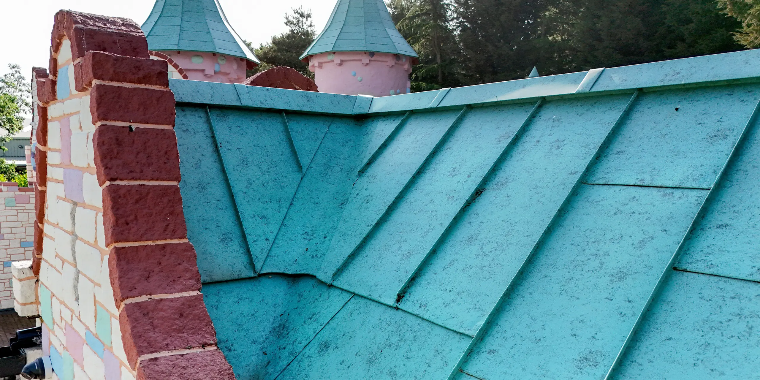 Gros plan sur une toiture à joints debout PREFALZ, couleur P.10 vert de gris, réalisée avec art à Sundown Adventureland. Elle impressionne par la précision de ses assemblages et la netteté de ses transitions. Les lignes de joints remarquables soulignent la forme dynamique de la toiture et créent un contraste visuel saisissant avec les pignons décoratifs colorés. L'aluminium PREFA démontre sa grande formabilité, notamment pour les géométries complexes, permettant des solutions précises. Par ailleurs, ce matériau se distingue par sa résistance à la corrosion, sa durabilité et sa protection fiable par tous les temps.
