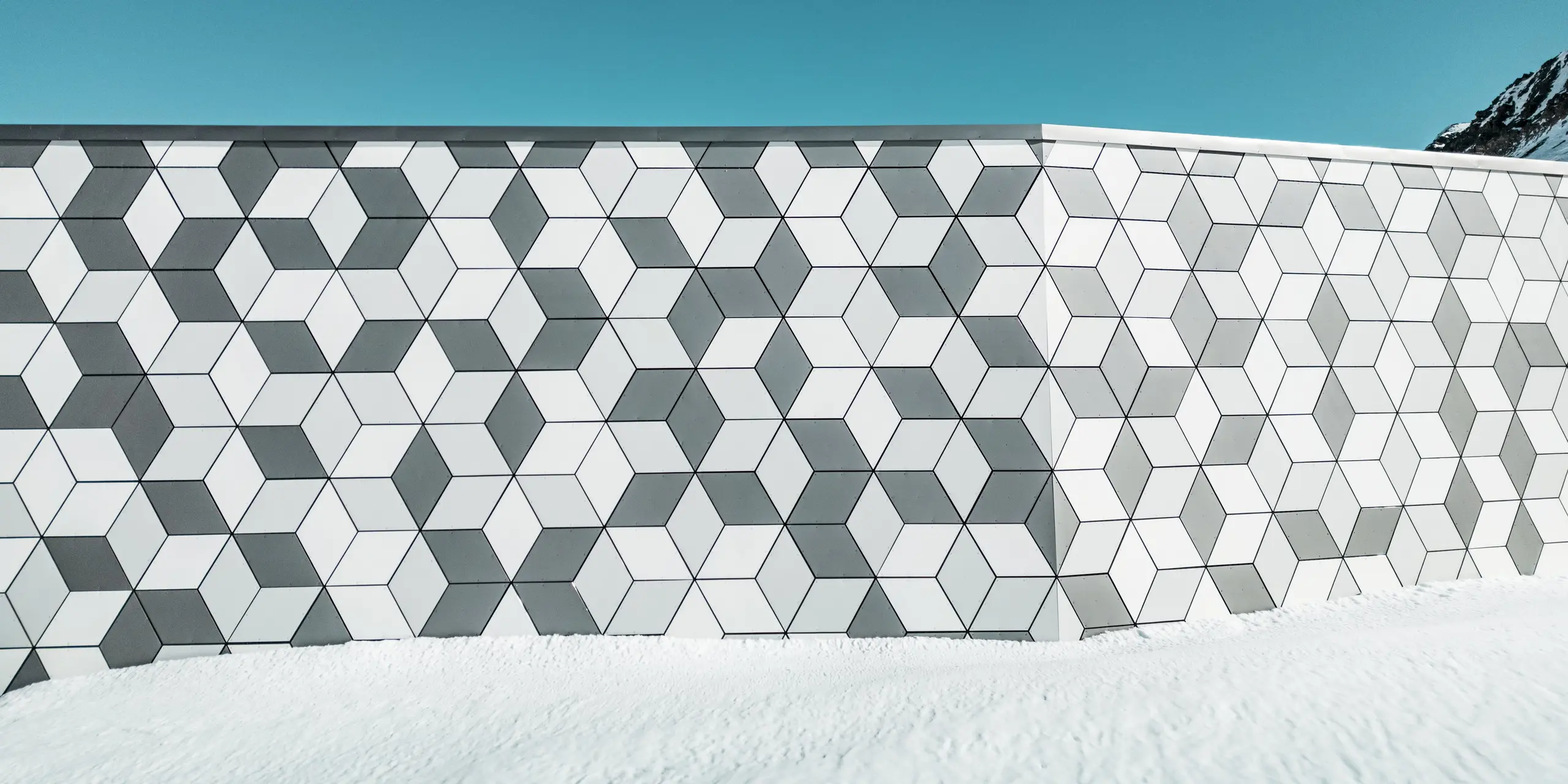 Ce gros plan de la façade de la station de téléphérique Jandri aux Deux Alpes illustre la disposition précise des losanges réalisés en panneaux composites en aluminium PREFABOND. Ces losanges forment un motif saisissant. Grâce à une palette de couleurs harmonieuse, déclinée en nuances de blanc et de gris, la façade s'intègre parfaitement au paysage alpin enneigé. Robustes et résistants aux intempéries, ces panneaux constituent une solution durable et esthétique pour les conditions climatiques extrêmes en montagne.