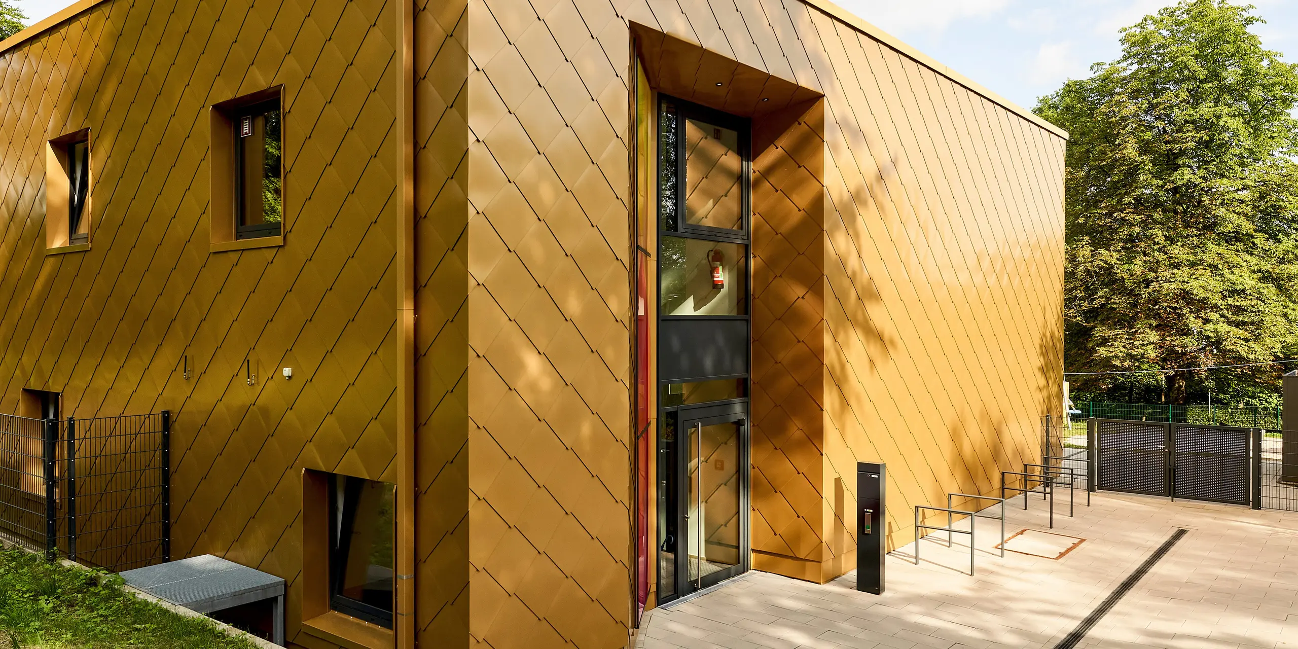 L'architecture cubique moderne, sublimée par une façade en aluminium PREFA au motif d'écailles dorées, attire immédiatement le regard. La précision de l'agencement des losanges crée un jeu d'ombres et de lumières saisissant. De généreuses surfaces vitrées et une entrée clairement délimitée soulignent la qualité du design. Durables et résistants à la corrosion, les produits en aluminium PREFA impressionnent par leur extrême robustesse, l'éclat de leurs couleurs et leur absence d'entretien – des solutions idéales pour des façades esthétiques et durables, parfaitement intégrées à la nature.