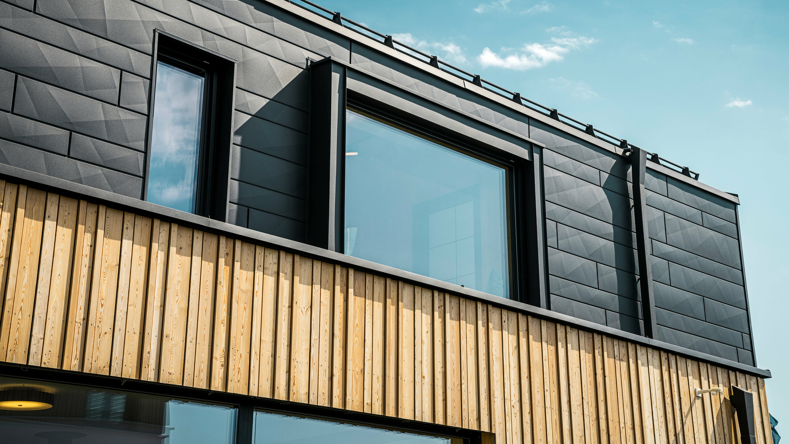Gros plan de l'étage supérieur de la maison témoin Autark dans le centre de maisons préfabriquées Blaue Lagune, Vösendorf (AT). La façade est constituée de panneaux en aluminium PREFA Siding.X en P.10 noir, qui se distinguent par leur aspect particulier avec des bords différents. En dessous, un élégant revêtement en bois forme un contraste harmonieux avec l'aluminium foncé. Les grandes fenêtres laissent entrer beaucoup de lumière à l'intérieur du bâtiment et soulignent le design moderne.