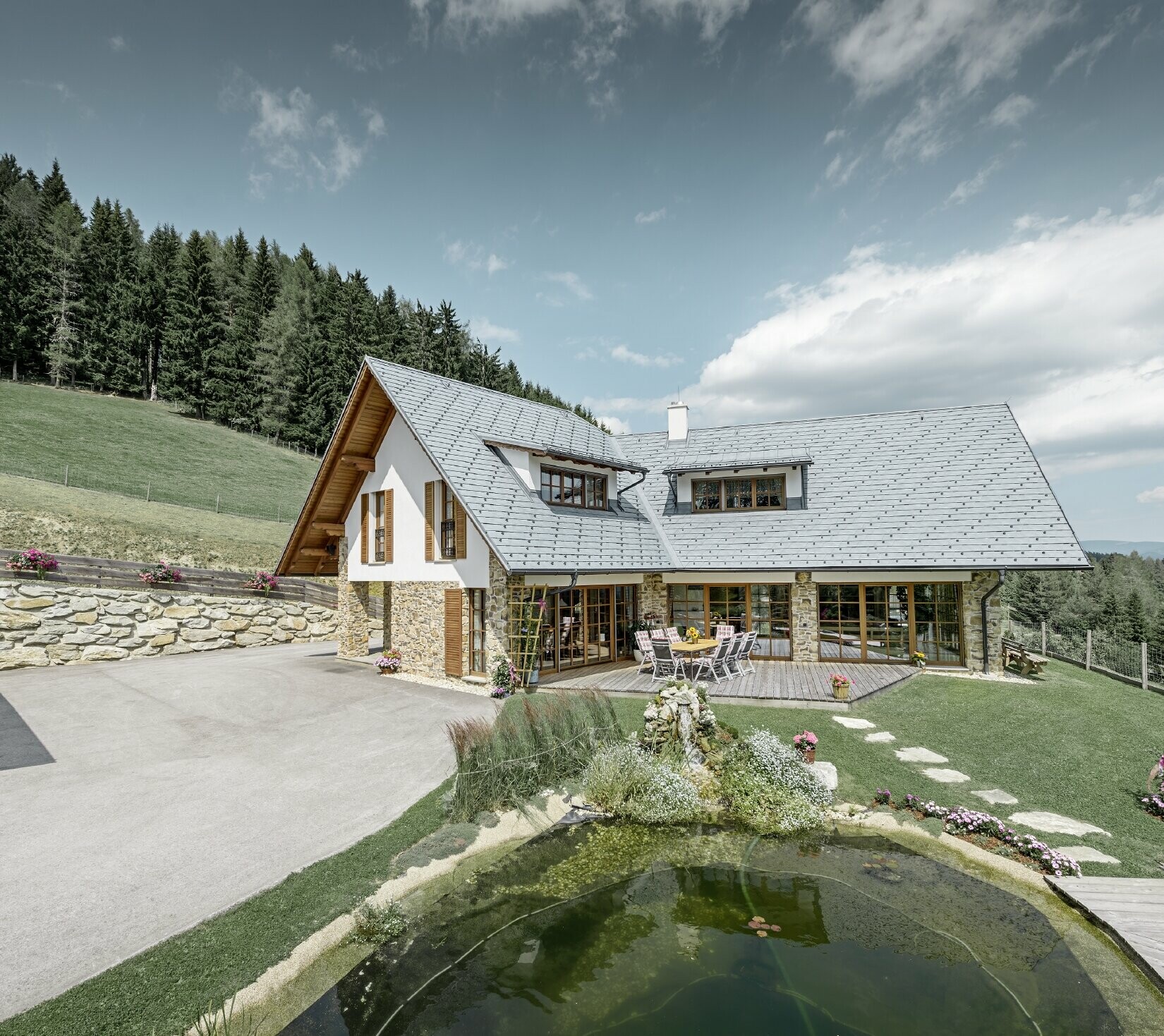 Construite en forme de L et à flanc de coteau, cette maison individuelle présente un toit à deux pans avec deux lucarnes rampantes. La toiture a été recouverte de bardeaux de toiture PREFA couleur gris pierre. Un étang se trouve au premier plan et on peut voir une forêt en arrière plan.