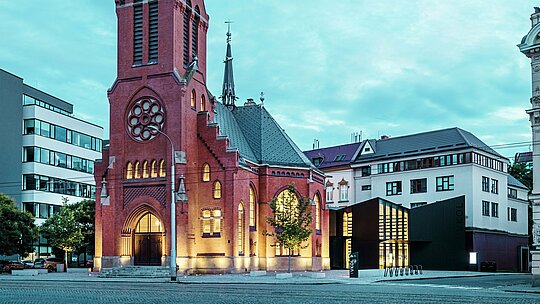 L'image illustre la fusion harmonieuse de l'architecture historique et moderne à Olomouc. L'imposante église rouge, avec sa flèche caractéristique et ses fenêtres gothiques, constitue le cœur historique de l'ensemble. Juste à côté s'élève le nouveau bâtiment moderne, dont la façade en panneaux composites en aluminium mat PREFABOND et en verre crée un contraste élégant avec l'architecture traditionnelle. Les larges façades vitrées offrent des aperçus sur le nouveau bâtiment, relié de manière innovante à l'église. Par sa forme précise et ses lignes épurées, la structure moderne contribue à l'atmosphère contemporaine sans compromettre le caractère historique de l'édifice. Cette symbiose réussie démontre comment des matériaux de haute qualité comme PREFABOND constituent le socle idéal pour la fusion de la tradition et de la modernité.