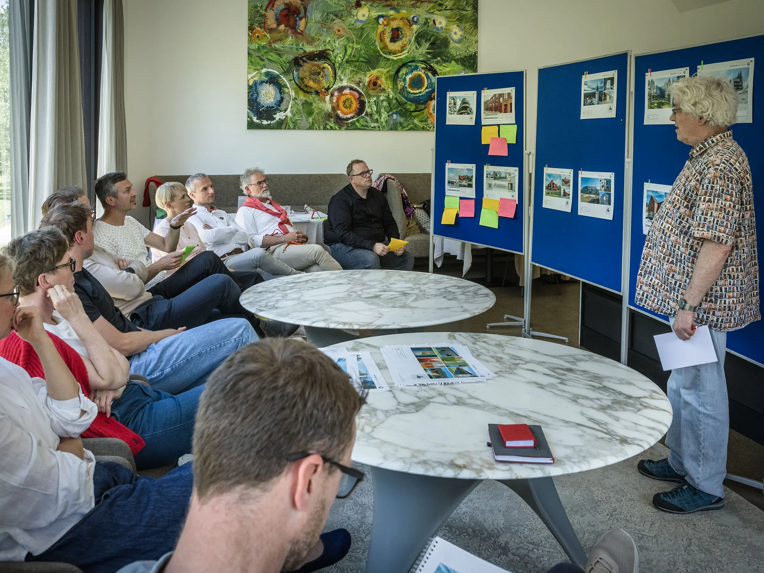 Réunion du jury de PREFARENZEN 2027 dans une salle de séminaire lumineuse : Matthias Finkentey, modérateur, présente des projets architecturaux en aluminium PREFA devant des tableaux d’affichage bleus, tandis que le jury, attentif, prend des notes. De larges fenêtres laissent entrer la lumière naturelle et offrent une vue sur un jardin verdoyant. Des livres d’architecture, des carnets et des stylos sont disposés sur les tables. L’atmosphère reflète une analyse approfondie, des échanges créatifs et une évaluation du design et de la fonctionnalité. Les produits en aluminium PREFA symbolisent des toitures et des façades durables et innovantes, alliant esthétique, qualité et inspiration pour les projets architecturaux.