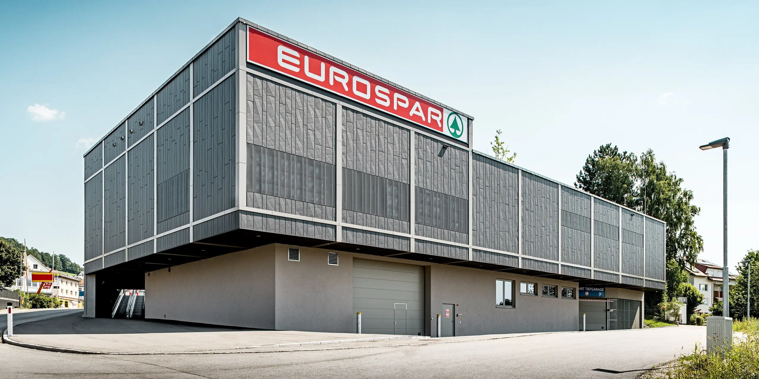 L'arrière du supermarché Eurospar de Rohrbach, y compris l'entrée du parking souterrain, présente une façade moderne en aluminium PREFA composée de panneaux Siding.X anthracite. La structure géométrique précise de ces panneaux lui confère une allure contemporaine et saisissante. Les panneaux perforés de la partie supérieure, particulièrement remarquables, offrent une texture sophistiquée tout en permettant une transmission lumineuse maîtrisée. Cette conception de façade confère au bâtiment une enveloppe durable, nécessitant peu d'entretien et esthétique, qui s'intègre harmonieusement à son environnement.