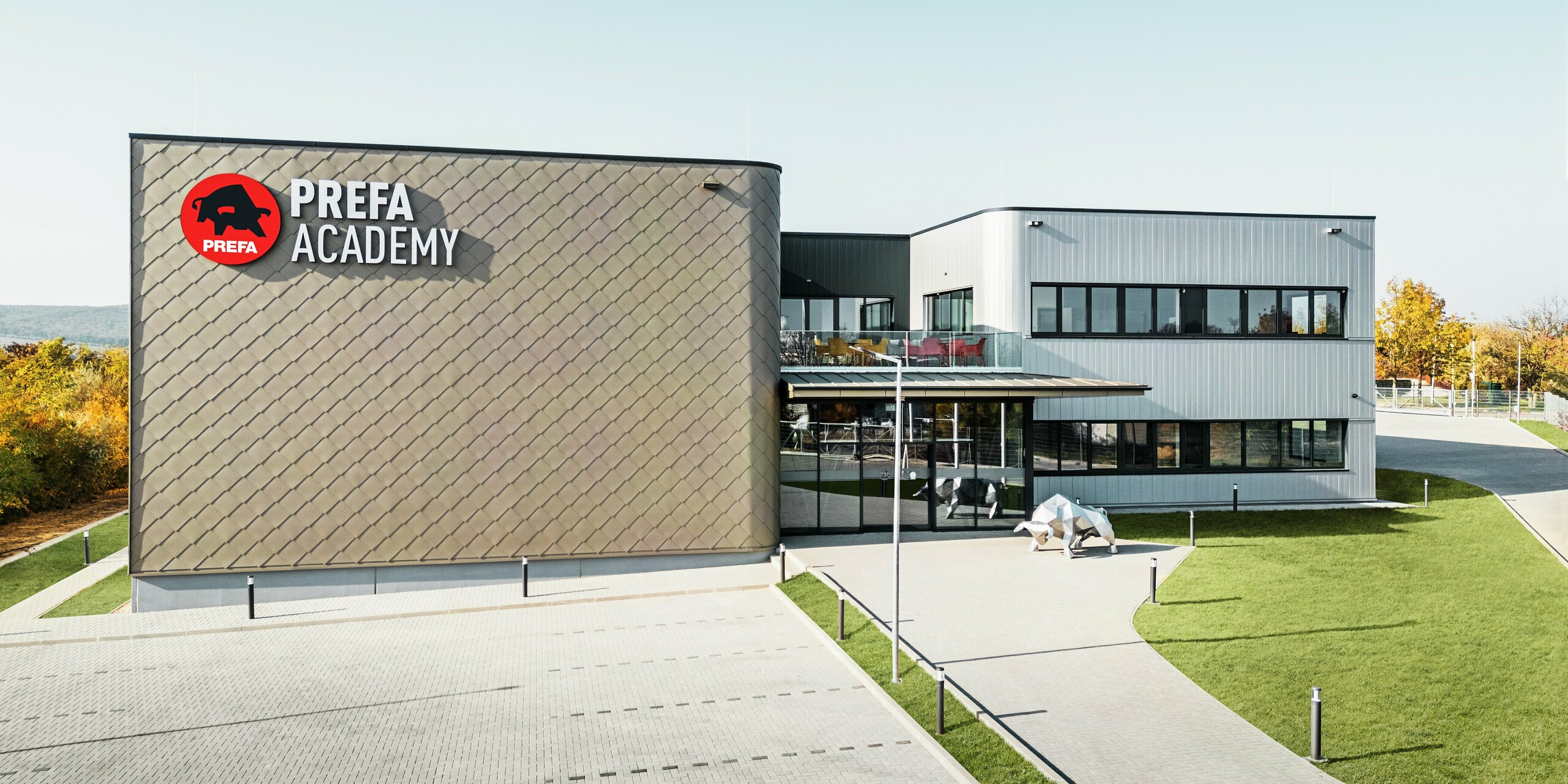 Le nouveau site de PREFA Hongrie à Törökbálint impressionne par sa façade moderne composée de losanges de façade PREFA 44 × 44 en bronze et de Sidings PREFA en argent métallisé. Les produits en aluminium durables garantissent une enveloppe de bâtiment résistante aux intempéries, facile d'entretien et visuellement attrayante. Le logo PREFA frappant et la sculpture du taureau en aluminium soulignent la puissance d'innovation, la qualité et la force de l'entreprise.