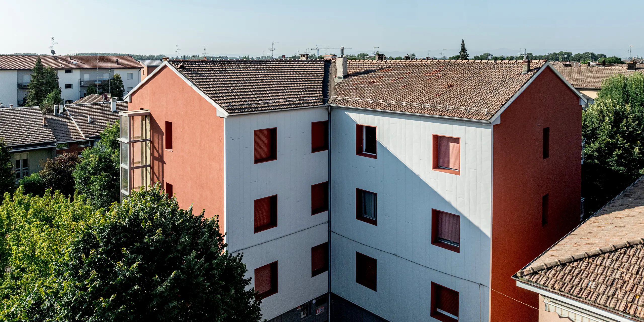 Vue du complexe résidentiel Alba B rénové, avec ses bâtiments à l'architecture affirmée et ses toits en pente, niché dans un quartier résidentiel établi et verdoyant. La façade, revêtue de panneaux d'aluminium PREFA Siding.X, se distingue par sa surface texturée et lumineuse, tandis que les encadrements de fenêtres d'un rouge éclatant apportent une touche d'originalité. La lumière naturelle crée de subtils jeux d'ombre et de lumière sur les panneaux, soulignant leur verticalité. La façade en aluminium PREFA se caractérise par sa durabilité, sa résistance aux intempéries et sa facilité d'entretien.