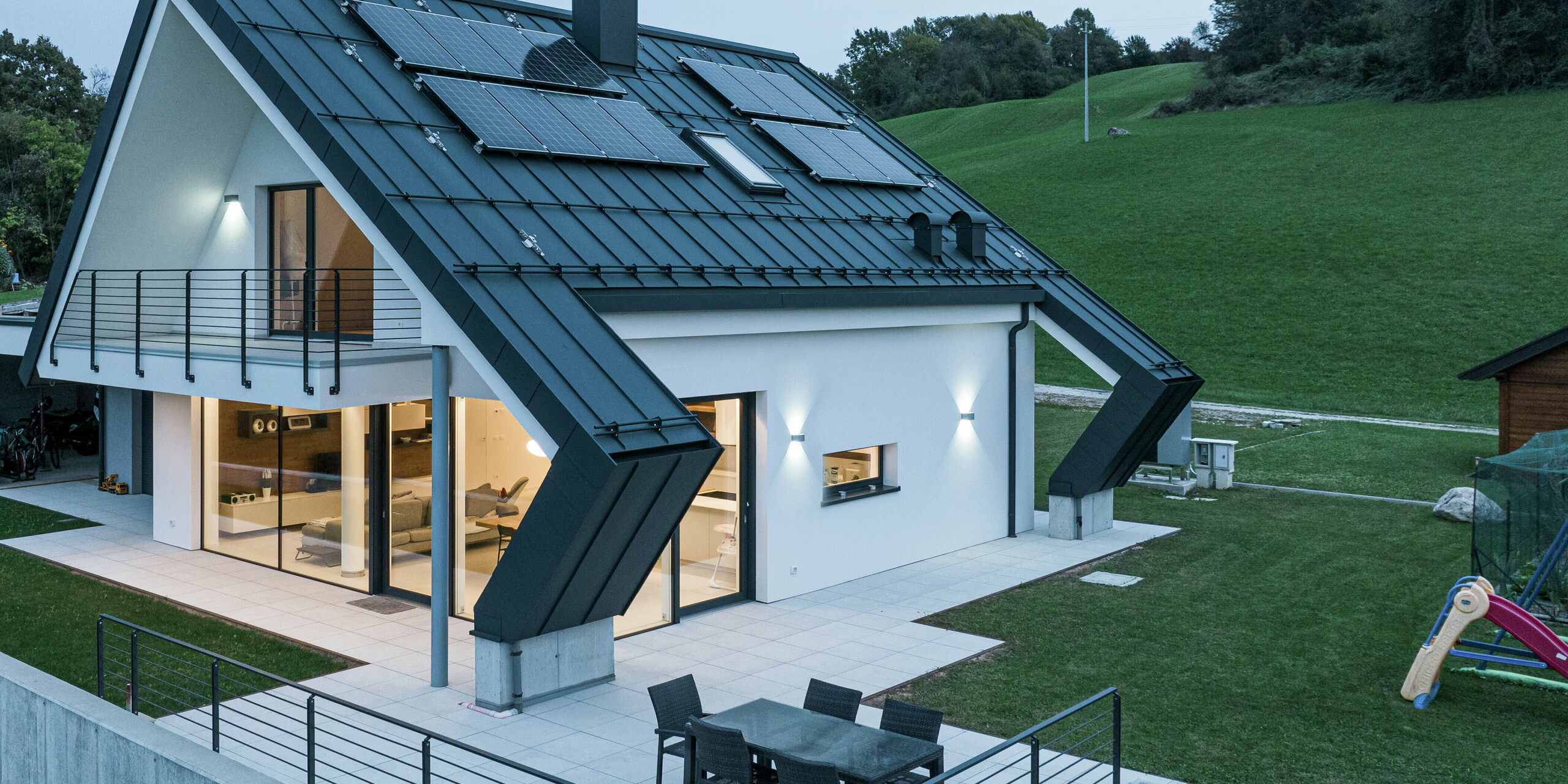 Maison individuelle moderne à Borgo Valbelluna avec toiture à joint debout PREFALZ en P.10 anthracite. Le toit en aluminium frappant et les modules photovoltaïques montés dessus allient esthétique et durabilité. Les surfaces de toit largement en surplomb offrent une protection supplémentaire à la façade et à la terrasse spacieuse. De grandes surfaces vitrées assurent une atmosphère de vie lumineuse et le balcon enveloppant souligne le design élégant.