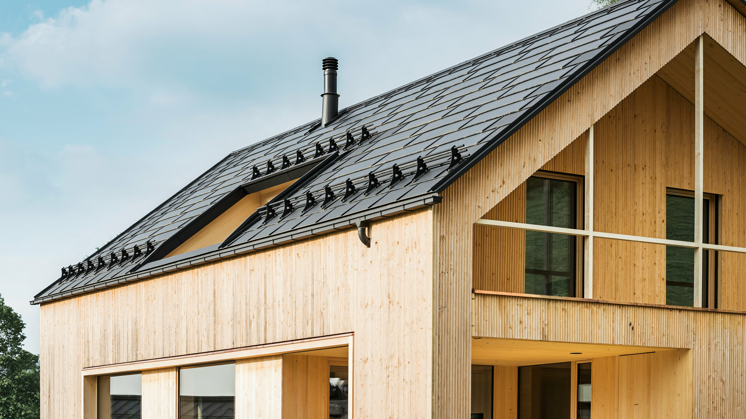 Gros plan sur le pignon d'une maison écologique à Egg, caractérisée par des tuiles solaires PREFA d'un noir élégant, qui s'intègrent harmonieusement au milieu des R.16 PREFA classiques. Les lignes épurées du toit contrastent avec la texture naturelle de la façade en bois, tandis que les tuiles solaires témoignent d'un mode de vie durable. La cheminée subtile s'intègre dans l'ensemble, qui allie un design respectueux de l'environnement et une esthétique rurale. La maison écologique innovante dispose également de deux balcons offrant une vue sur le magnifique paysage montagneux d'Egg dans le Vorarlberg.