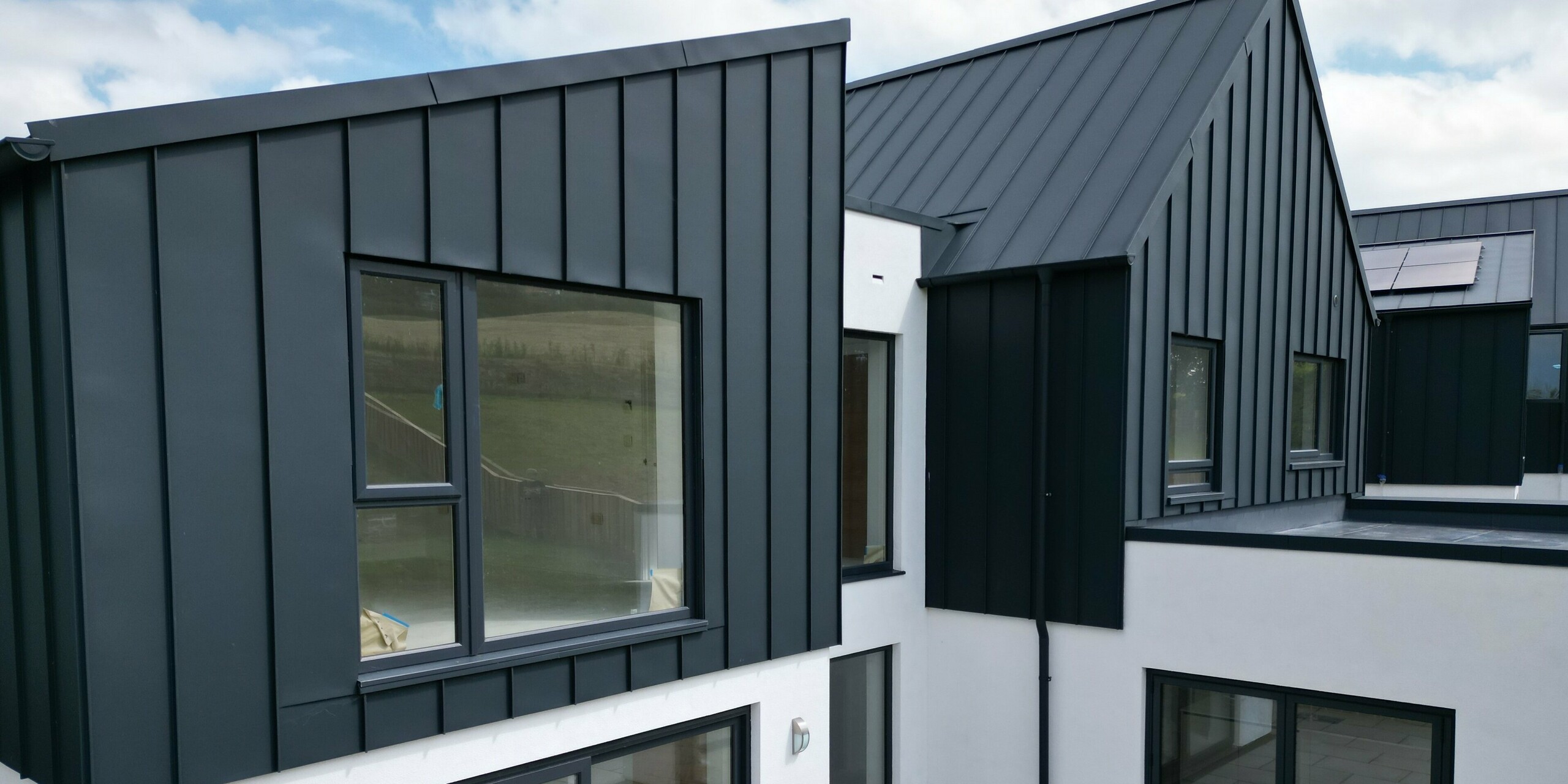 Vue détaillée d'une maison individuelle avec jardin et terrasse, recouverte de PREFALZ en P.10 anthracite. La toiture à joint debout en aluminium allie design moderne et durabilité et souligne l'approche de construction durable. Les grandes fenêtres apportent beaucoup de lumière à l'intérieur.