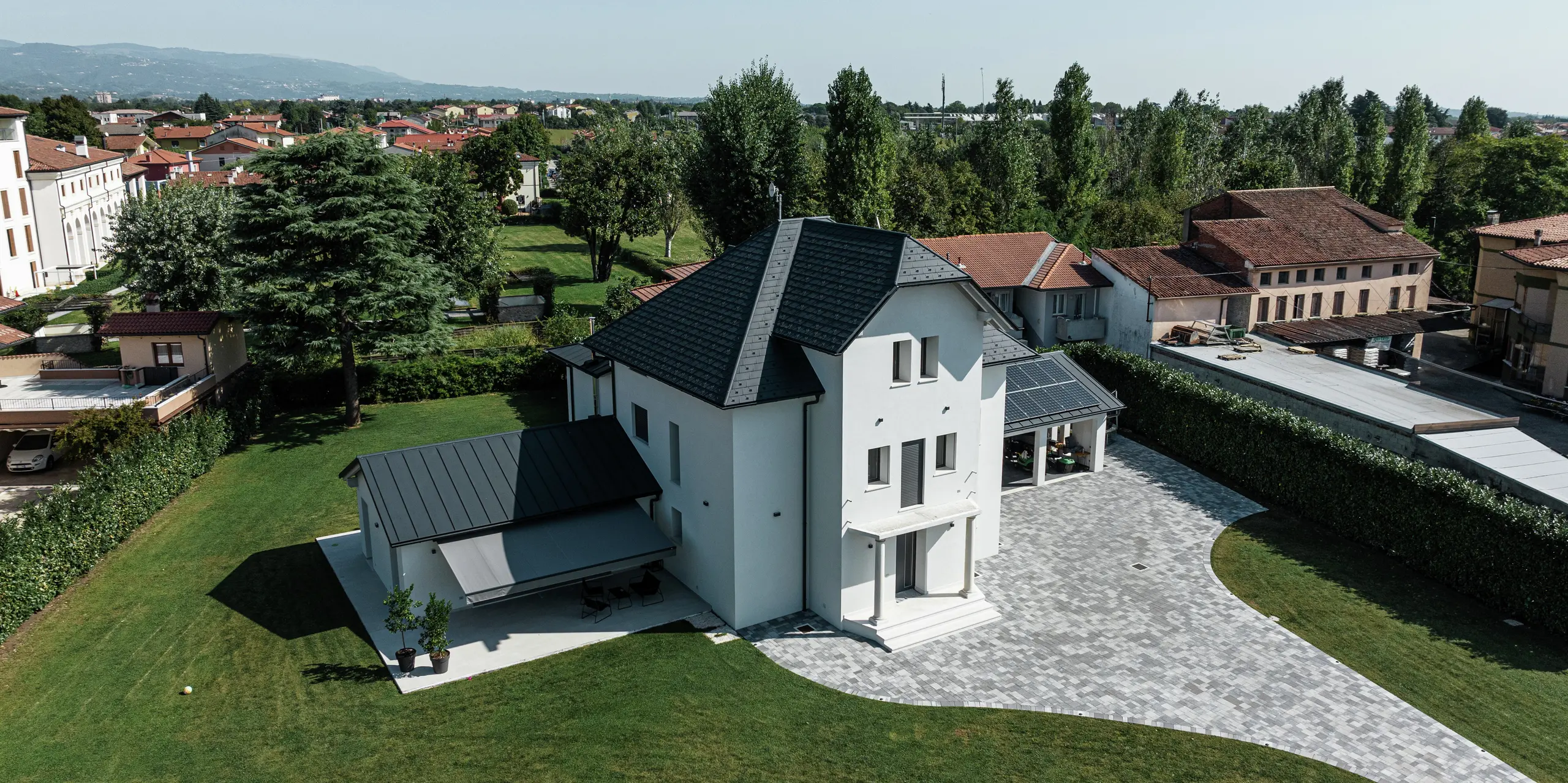 Vue aérienne d'une maison familiale moderne à Marano Vicentino, en Italie, avec des bardeaux de toiture PREFA durables en P.10 anthracite. Les dépendances adjacentes sont couvertes d'une toiture à joint debout en aluminium. La solution de toiture en aluminium de haute qualité allie esthétique, durabilité et résistance aux intempéries.