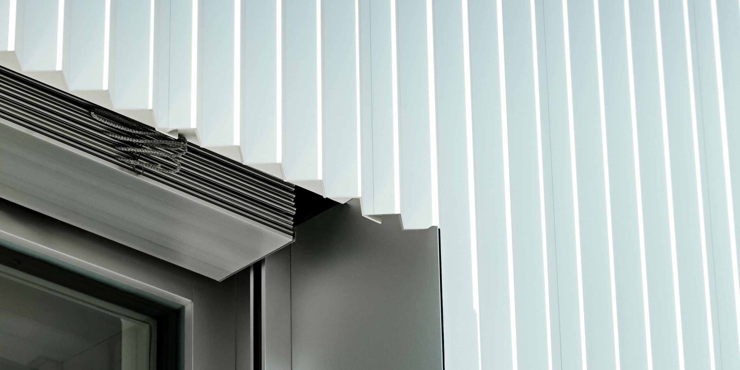 Gros plan précis d'un encadrement de fenêtre avec profilé triangle PREFA P.10 Blanc Pur. Les profilés en aluminium aux contours nets épousent parfaitement la fenêtre, complétée par des guides extérieurs intégrés pour stores vénitiens. La finition haut de gamme témoigne du savoir-faire irréprochable, de la durabilité et de la résistance aux intempéries des systèmes de façade PREFA – idéaux pour une architecture moderne et raffinée.