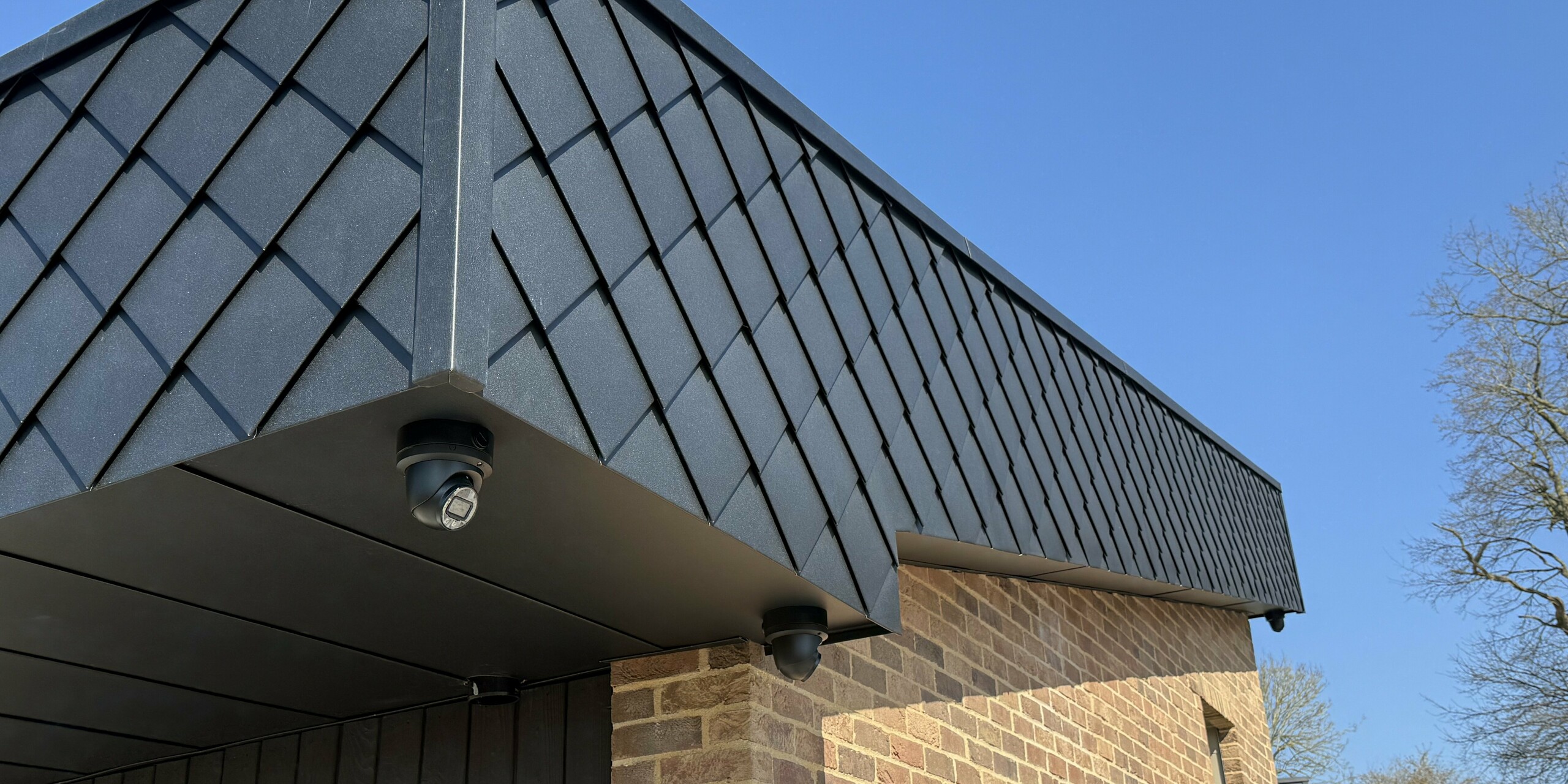 Vue détaillée d'une façade moderne du projet résidentiel « Doone Brae Farm », caractérisée par une transition saisissante entre la brique clinker chaleureuse et les élégants losanges de façade PREFA 20 × 20 en P.10 anthracite. Ces panneaux en aluminium créent un effet visuel dynamique et soulignent la pureté des lignes du bâtiment. Le contraste entre la brique clinker naturelle et l'aluminium de haute qualité confère au design un caractère intemporel et une grande efficacité énergétique.