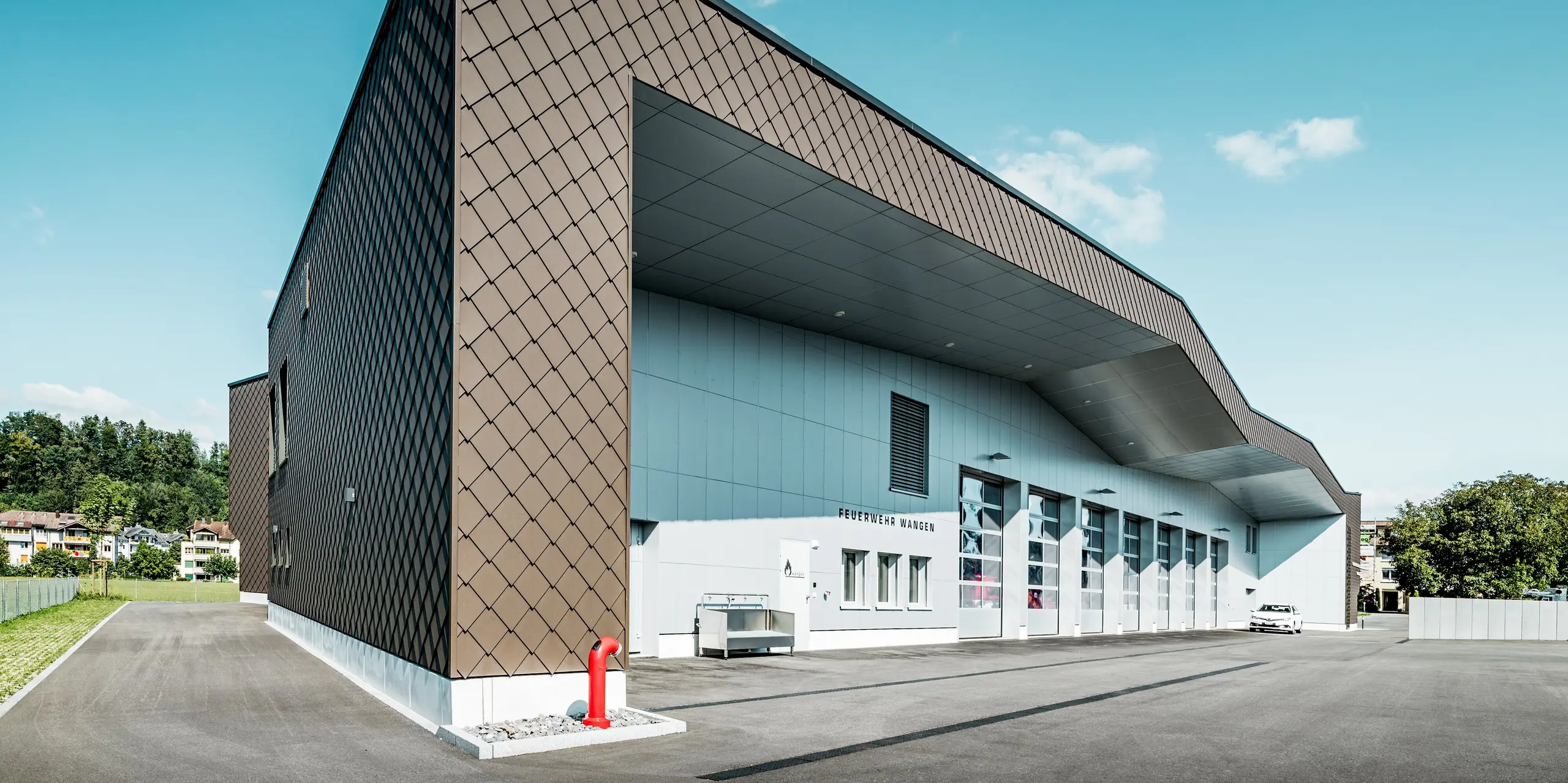 Vue d'ensemble impressionnante de la caserne de pompiers et de la centrale électrique de Wangen, en Suisse. La façade à grande échelle, composée de losanges PREFA 29 x 29 en P.10 Brun, crée une profondeur exceptionnelle grâce à son motif en losange saisissant. Les panneaux composites en aluminium P.10 Prefa Blanc apportent des contrastes modernes. Durables, les produits en aluminium PREFA allient une architecture sophistiquée à une grande résistance aux intempéries, un entretien réduit et une valeur pérenne.
