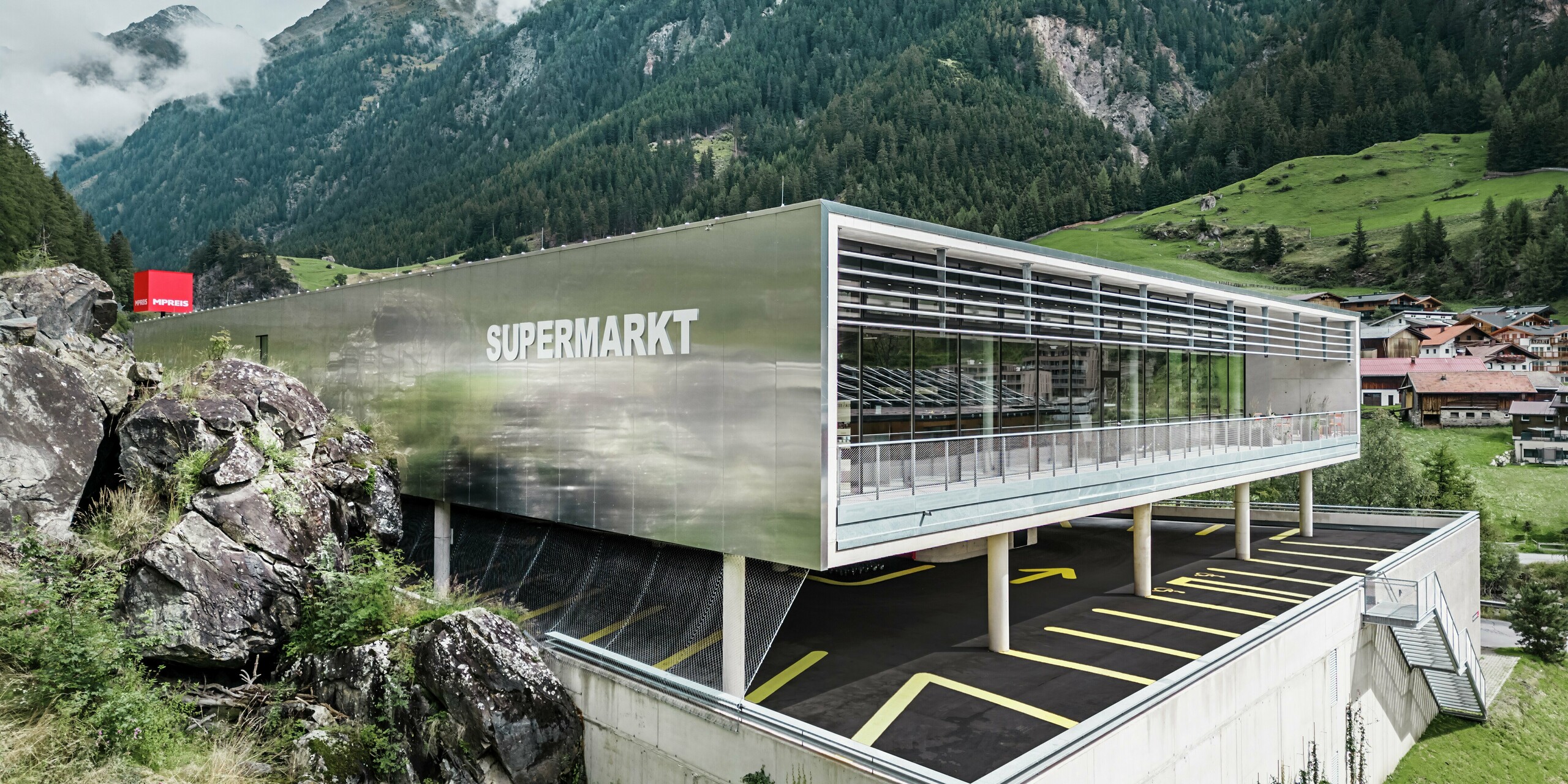 Supermarché MPREIS à Sölden, au Tyrol, avec des panneaux composites en aluminium dans la couleur spéciale Reflective Natural. La façade reflète le paysage montagneux environnant des Alpes de l'Ötztal et allie design moderne et nature. L'architecture claire avec le parking sous le supermarché crée une opportunité d'achat innovante pour la région alpine.