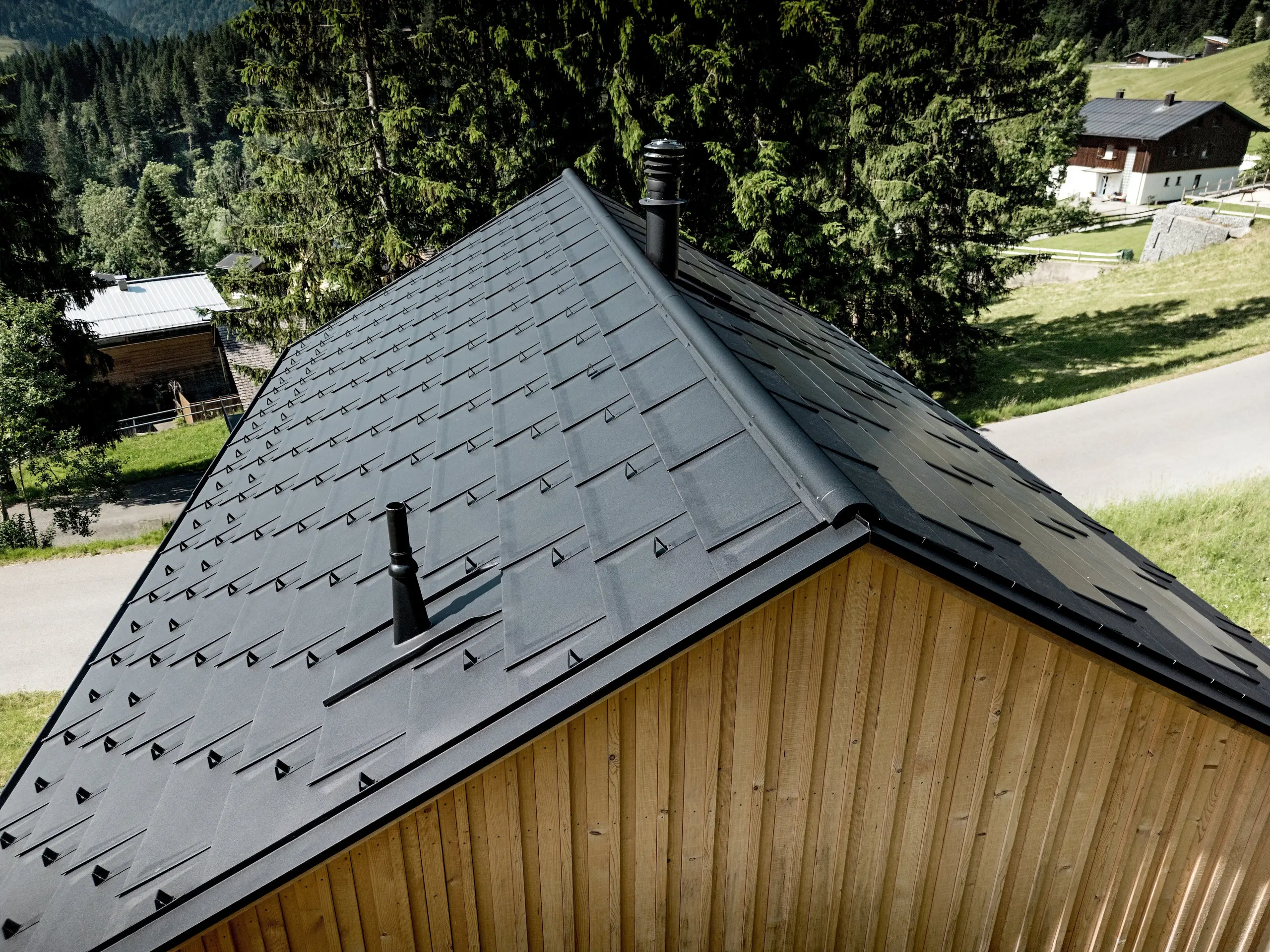 Cette vue rapprochée dynamique met en valeur l’imposante toiture de la maison Immi, sur fond de prairies verdoyantes et de sapins sombres. Les R.16 en P.10 noir, les détails de finition soignés et les arrêts-neige composent une toiture à l’esthétique puissante et homogène. Légère et robuste, la couverture en aluminium résiste aux conditions météorologiques alpines ainsi qu’aux fortes charges de neige, tout en protégeant durablement la maison en bois d’Ebnit avec un entretien minimal.