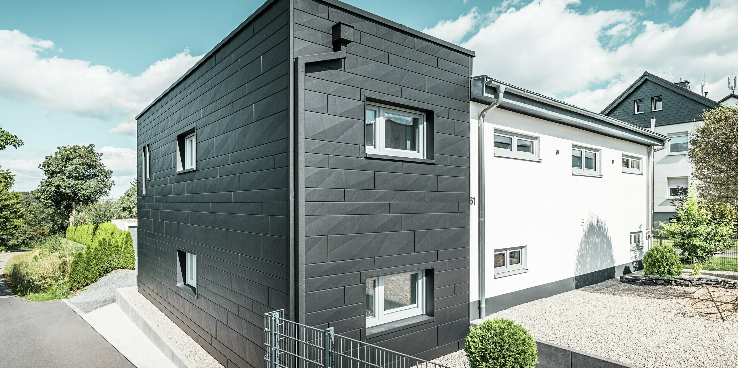La remarquable façade Siding.X PREFA en P.10 noir définit l'architecture moderne de cette maison individuelle rénovée à Teuschnitz. Les panneaux en aluminium à la structure claire confèrent au bâtiment une esthétique élégante et géométrique. Le système de gouttière PREFA intégré, avec son tuyau carré, s'intègre parfaitement au bâtiment, offrant une solution fonctionnelle et esthétique. Le jardin verdoyant et la zone de gravier devant la maison complètent le design minimaliste et soulignent le caractère moderne de la maison.