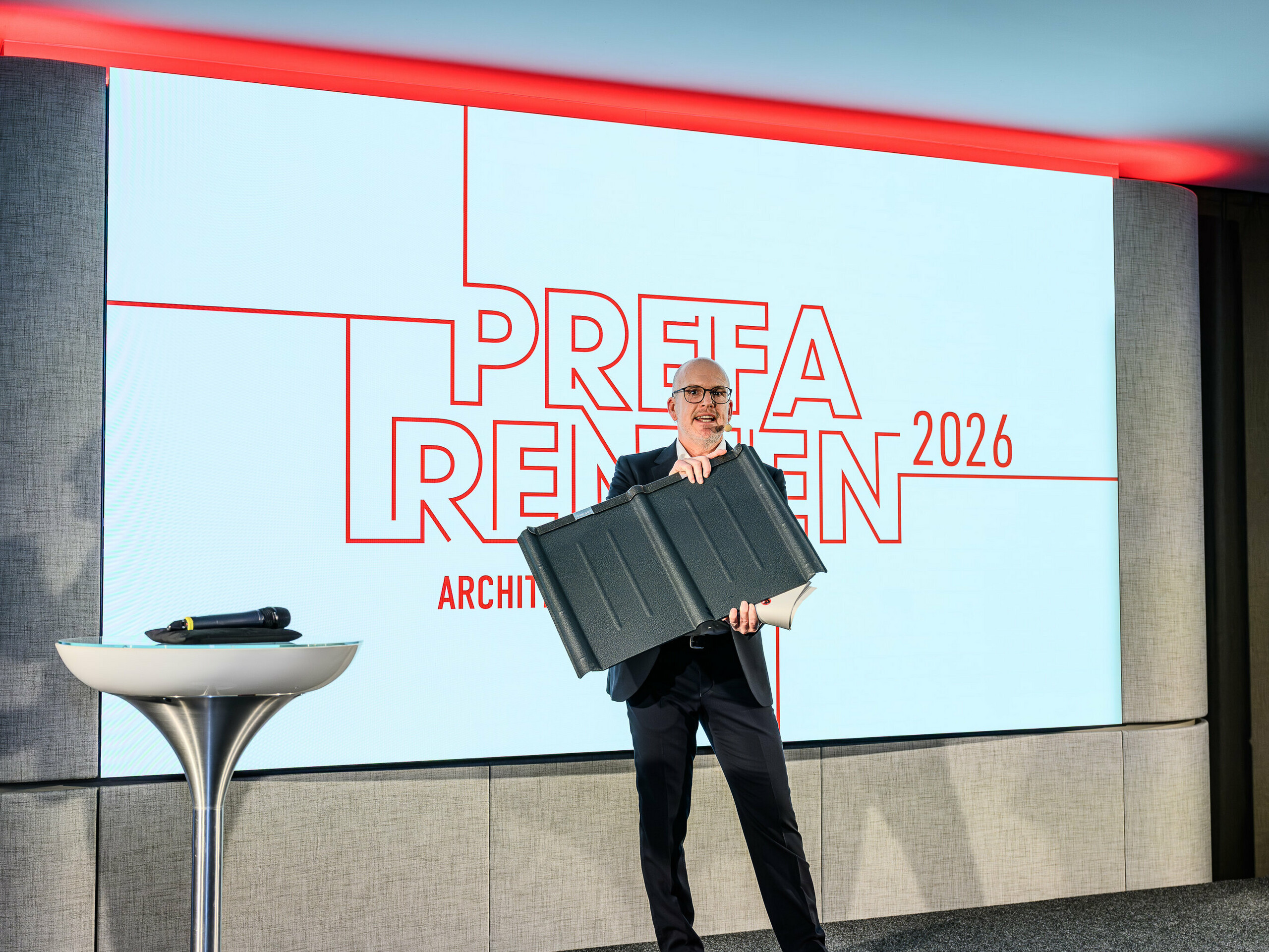 Moment fort de la présentation PREFARENZEN 2026 : Jürgen Jungmair, directeur marketing international de PREFA, présente avec fierté le panneau de toiture PREFA, une référence dans le domaine. L’accent est mis sur ce produit en aluminium durable, fabriqué avec succès depuis près de 80 ans. La scène moderne, ornée du logo PREFARENZEN, souligne l’importance de ce produit pour l’architecture d’avenir. Léger, résistant aux intempéries et inoxydable, le panneau de toiture PREFA allie technologie et esthétique à la perfection.