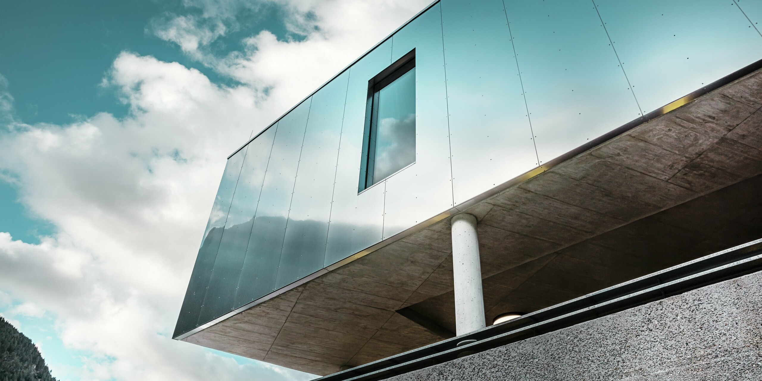 Architecture moderne du supermarché MPREIS à Sölden, au Tyrol, avec des panneaux composites en aluminium PREFA réfléchissants dans la couleur spéciale Reflective Natural. La façade brillante reflète le ciel et le paysage montagneux. Des lignes épurées et des colonnes massives en béton allient un design innovant à la stabilité et à l'élégance.