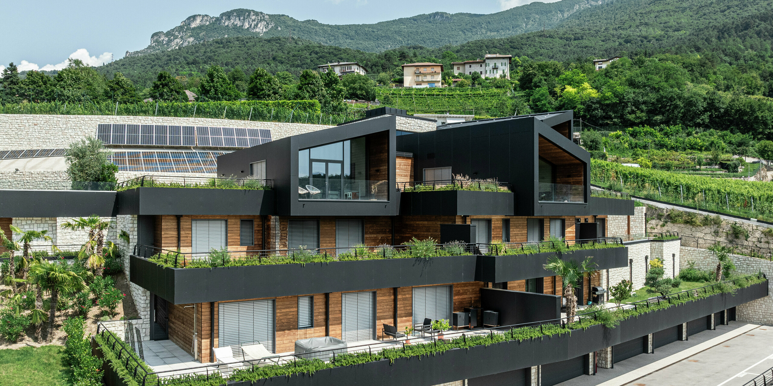 [Translate to Français:] Moderne Wohnanlage in Hanglage mit nachhaltigem Design: Die Kombination aus Naturstein, Holz und der hochwertigen PREFA Aluminiumfassade in P.10 Schwarz verleiht dem Gebäude eine elegante und zeitlose Optik. Das PREFALZ Dach in P.10 Schwarz bietet optimalen Schutz und Langlebigkeit. Großzügige Terrassen, begrünte Dächer und Photovoltaikanlagen unterstreichen das nachhaltige Wohnkonzept. Die harmonische Einbettung in die Landschaft sowie die hochwertigen und pflegeleichten PREFA Materialien garantieren eine stilvolle und witterungsbeständige Architektur mit höchstem Wohnkomfort.