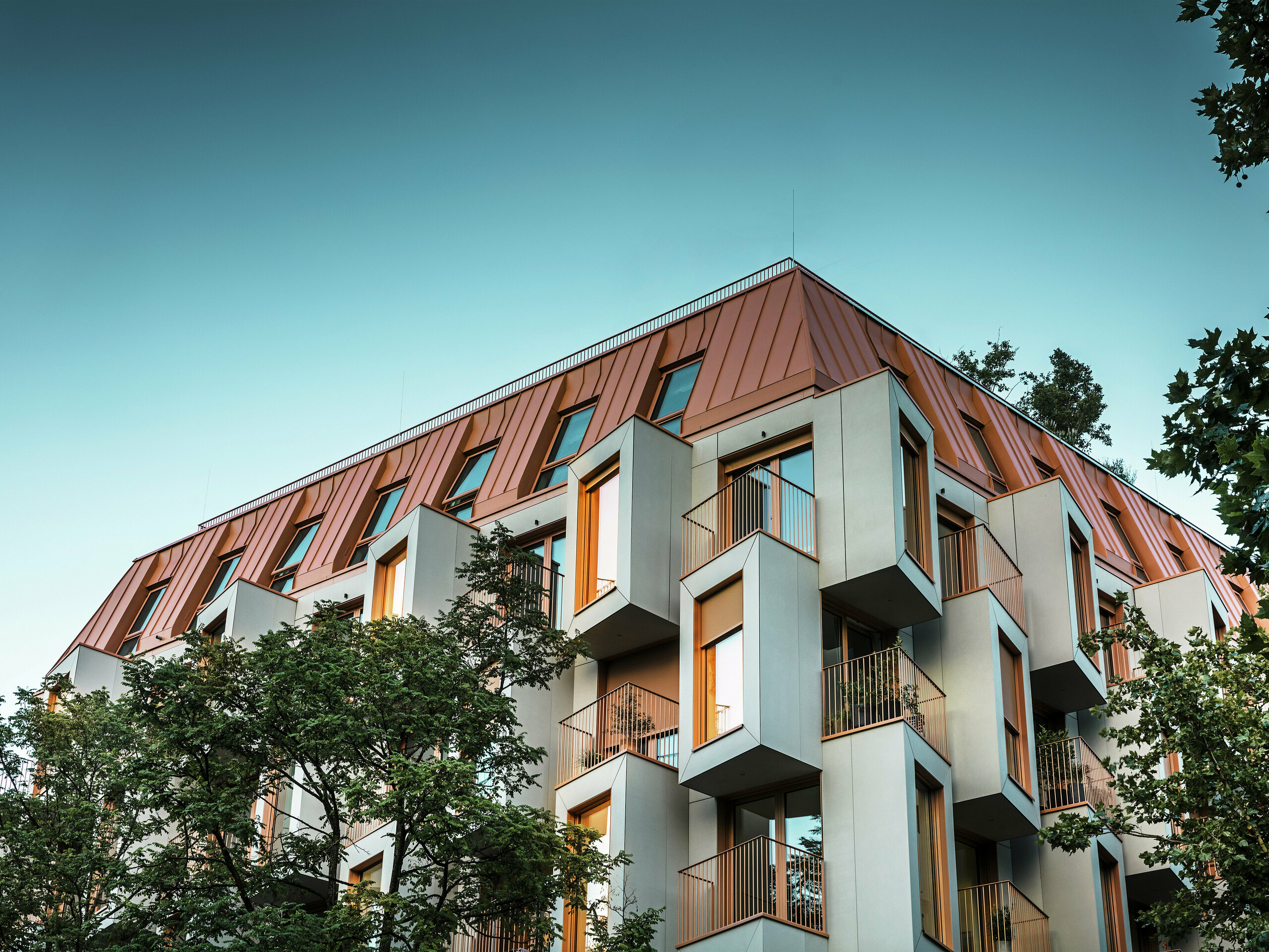 Façade architecturalement remarquable du projet résidentiel Van B à Munich avec des baies vitrées remarquables et un toit en aluminium PREFALZ dans la couleur spéciale IGP Orange HDP. Au crépuscule, la vue d'en bas montre les lumières dans les fenêtres. La solution de toiture moderne combine une résistance maximale aux intempéries avec une apparence élégante et apporte des accents colorés dans l'espace urbain. L'interaction du béton renforcé de fibres de verre, des cadres de fenêtres couleur cuivre-orange et du toit en aluminium crée une enveloppe de bâtiment dynamique, durable et durable - idéale pour une construction tournée vers l'avenir.