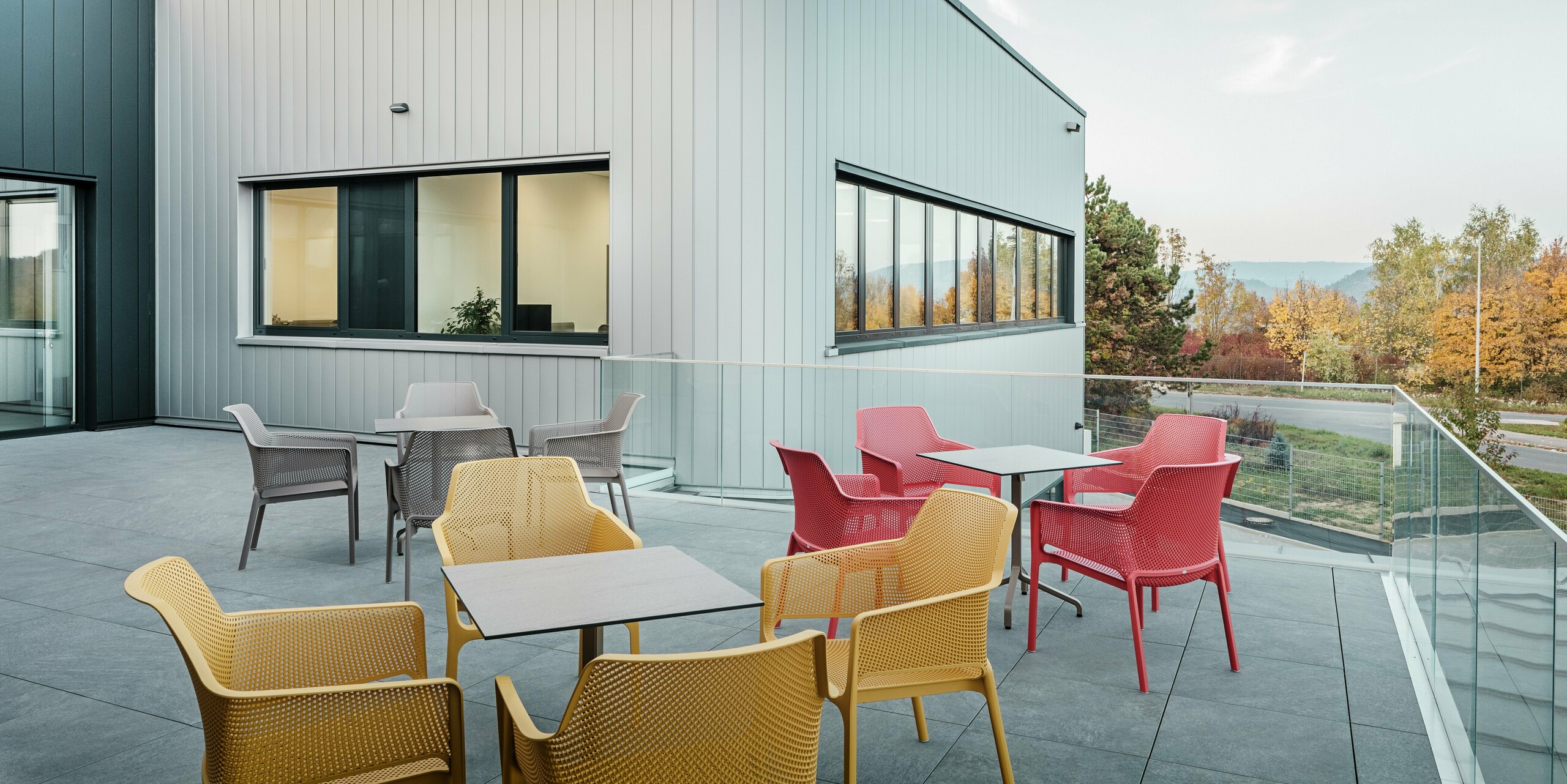 La terrasse moderne du nouveau site PREFA à Törökbálint offre un espace extérieur accueillant avec vue sur le paysage automnal. La façade est revêtue de Sidings PREFA en argent métallisé et anthracite, qui se caractérisent par leur aspect élégant, leur grande résistance aux intempéries et leur entretien facile. La finition précise et la conception droite et verticale soulignent le design contemporain du bâtiment. Les sièges colorés au premier plan ajoutent des accents colorés au décor automnal.