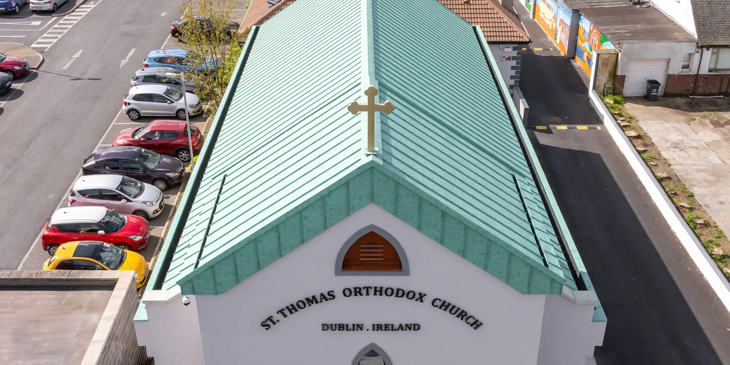 Prise de vue par drone de la vue de face de l'église orthodoxe Saint-Thomas de Dublin avec son remarquable toit à joints debout en aluminium PREFALZ de couleur P.10 vert de gris. La croix visible de loin sur le pignon, les élégantes fenêtres cintrées et la façade clairement structurée confèrent à l'édifice sacré une présence incomparable. Le grand paysage de toiture avec des lignes de joint debout précises et l'aspect patiné caractéristique rappelle les toits en cuivre traditionnels et met un fort accent architectural dans l'environnement urbain. L'aluminium PREFA de haute qualité impressionne par sa résistance à la corrosion, son faible poids, sa longévité et sa qualité de couleur durable - idéal pour une architecture sophistiquée et des solutions de toiture durables.