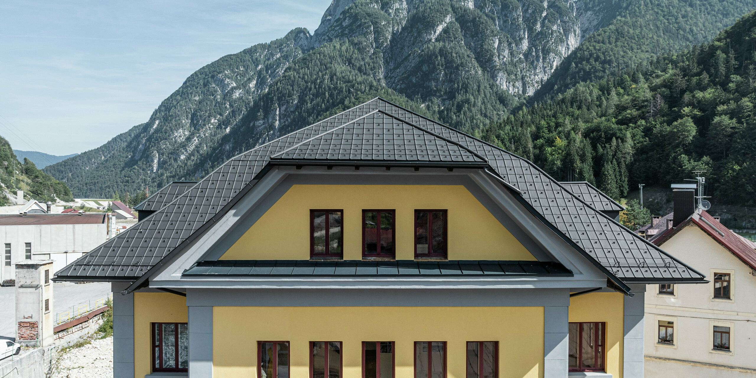 Gros plan de la couverture du Museo Cave del Predil en Italie. Le bâtiment jaune du musée a été recouvert de tuiles PREFA en P.10 anthracite. La solution de toiture robuste en aluminium s'intègre harmonieusement au bâtiment historique avec sa façade jaune. À l'arrière-plan se trouve l'impressionnant paysage alpin avec des pentes boisées, ce qui exige que les produits PREFA soient extrêmement résistants aux intempéries.