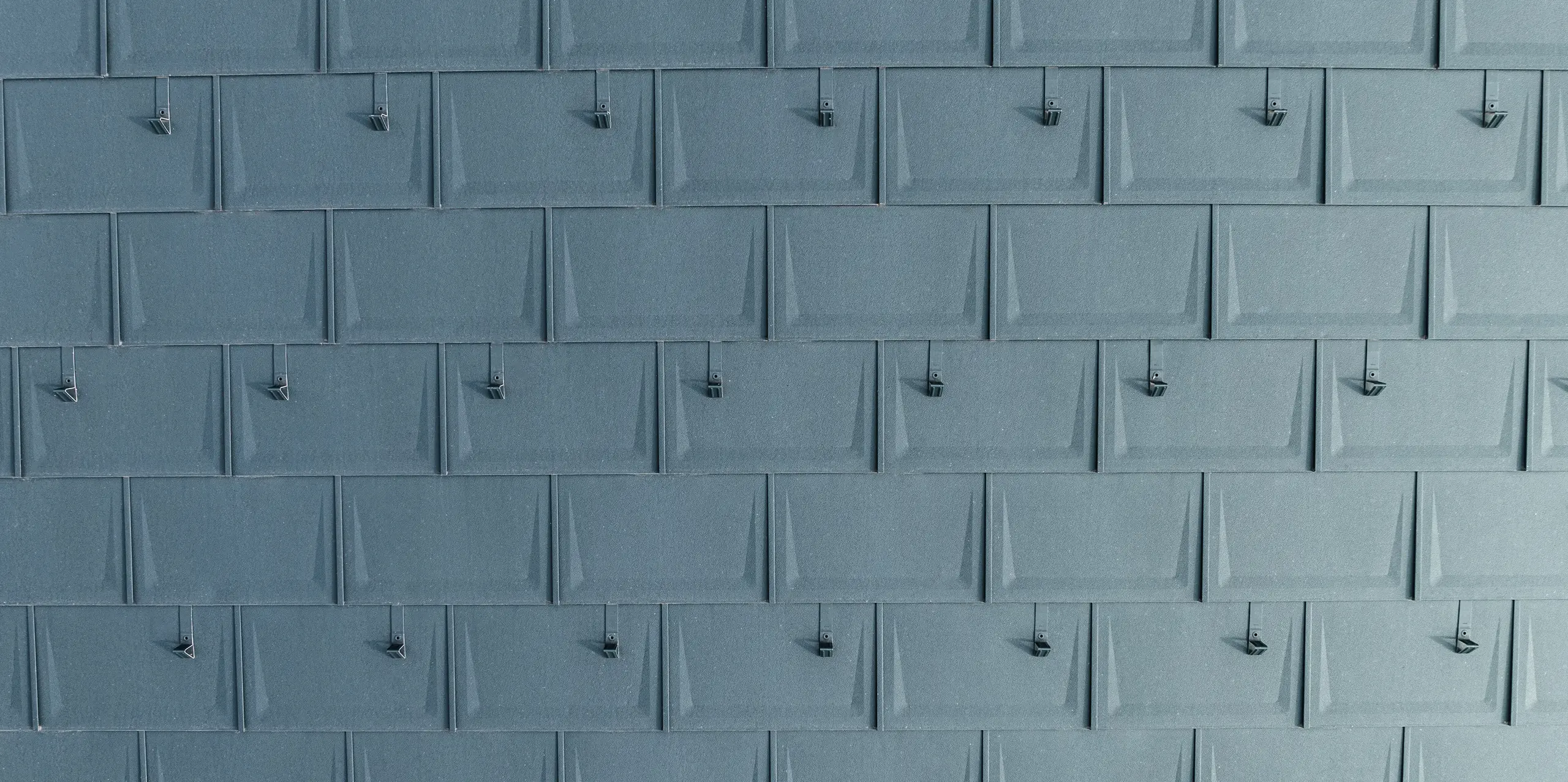 Vue détaillée des PREFA R.16 posés avec précision en P.10 anthracite avec arrêts de neige montés. Le panneau de toit linéaire en aluminium impressionne par son aspect moderne, sa durabilité et sa résistance extrême aux intempéries. Grâce à une technologie de fixation sophistiquée, il est particulièrement résistant aux tempêtes et aux cassures. Les butées à neige offrent une sécurité supplémentaire dans les régions enneigées et empêchent les masses de neige de glisser de manière incontrôlable.