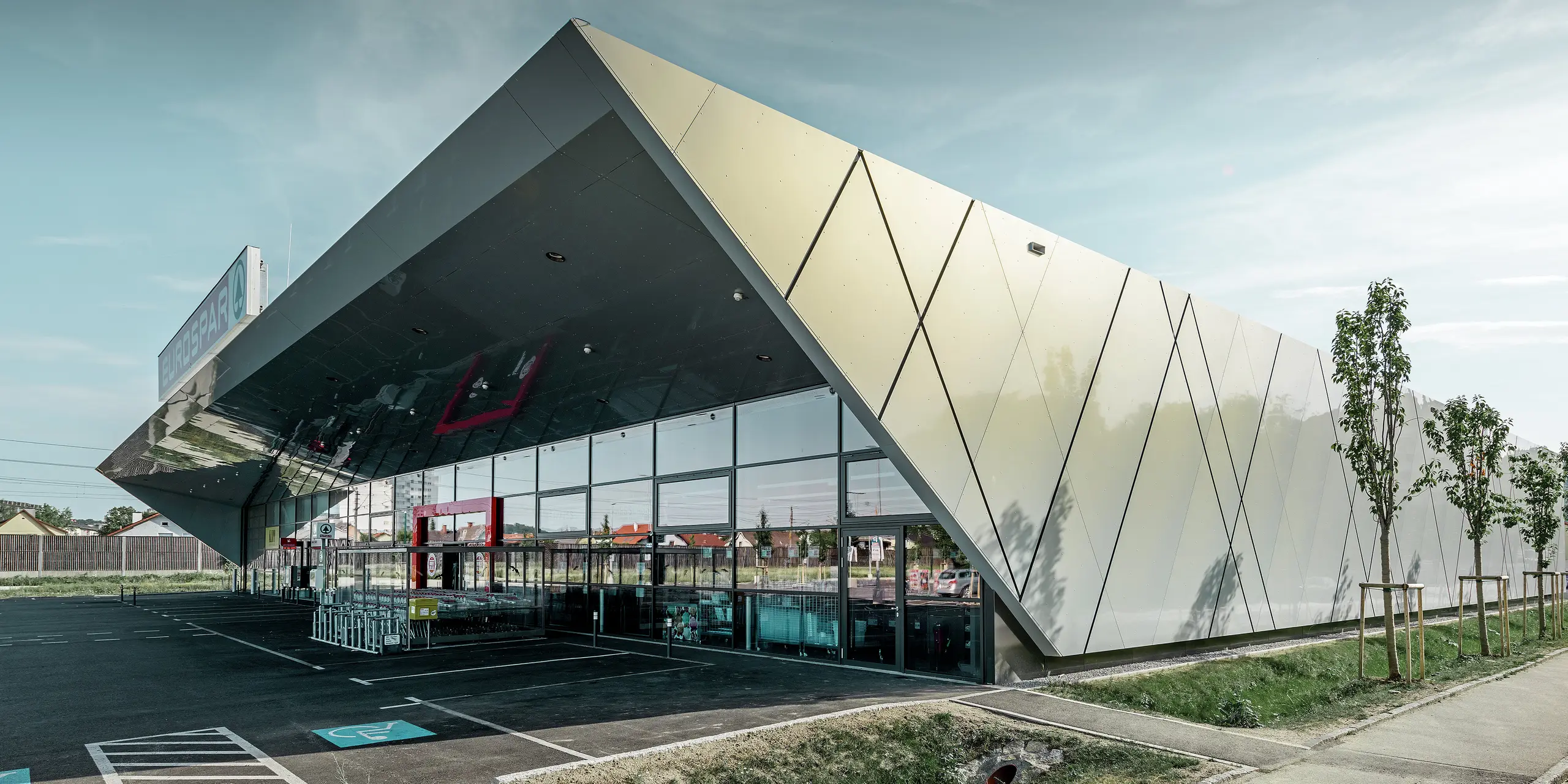 Entrée de la nouvelle filiale Eurospar de Wels habillée de panneaux de façade modernes de PREFA couleur argent doré ; selon l’angle de vue, à la lumière du soleil les panneaux composites en aluminium chatoient dans différentes couleurs.