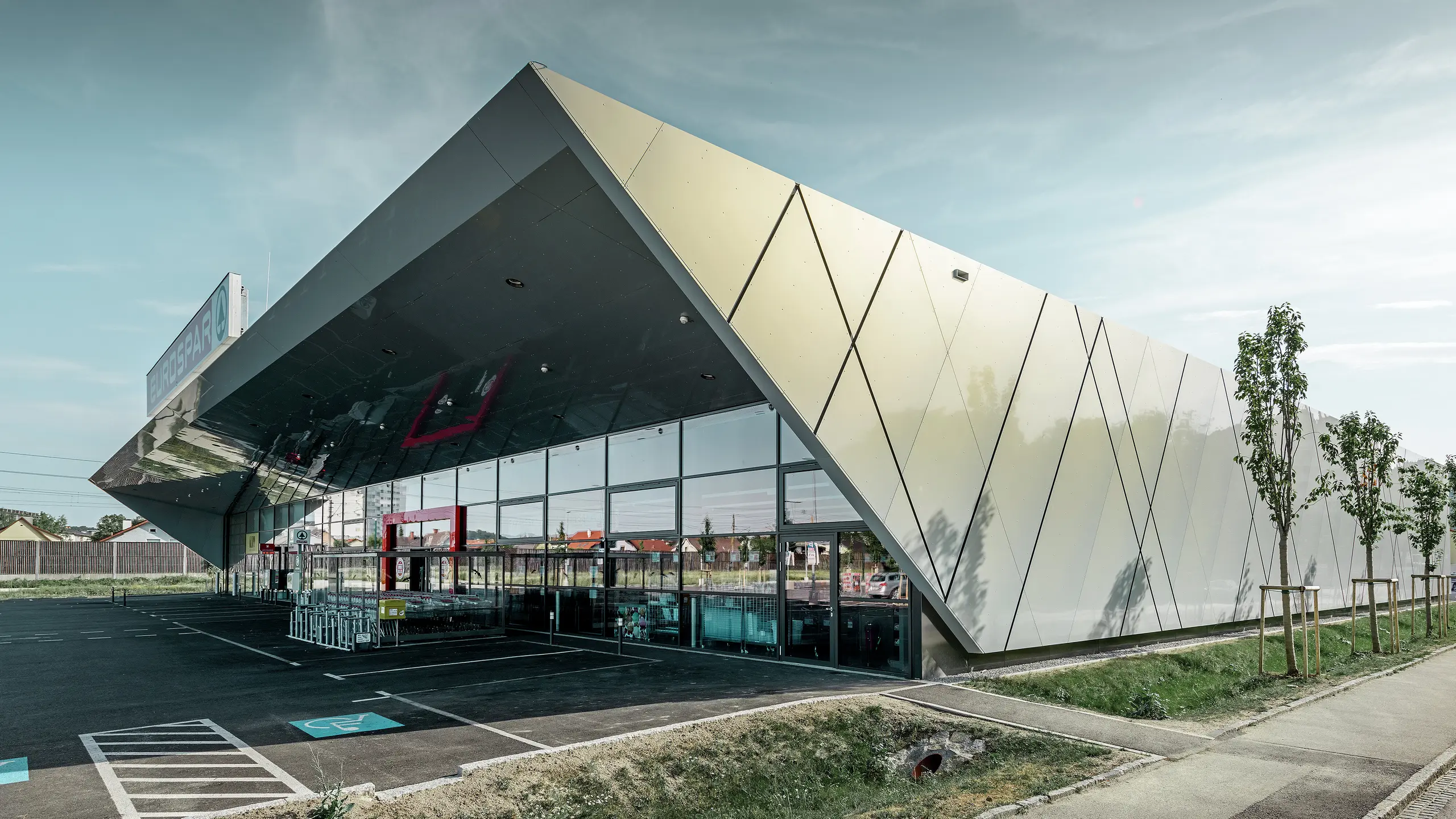 Entrée de la nouvelle filiale Eurospar de Wels habillée de panneaux de façade modernes de PREFA couleur argent doré ; selon l’angle de vue, à la lumière du soleil les panneaux composites en aluminium chatoient dans différentes couleurs.