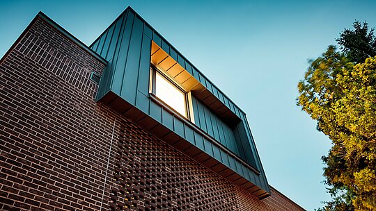 La photographie présente la façade moderne d'une villa multigénérationnelle à Budapest, vue sous un angle dynamique et en contre-plongée. Le cube en porte-à-faux, revêtu d'aluminium PREFALZ P.10 anthracite, contraste de façon saisissante avec les briques rouge foncé en contrebas. La précision de l'exécution des joints verticaux souligne la pureté des lignes et met en valeur la haute qualité des produits en aluminium PREFA. La lumière chaude crée un jeu d'ombres et de lumière saisissant entre le métal, la brique et l'ombre, révélant avec élégance le caractère novateur et intemporel du bâtiment.