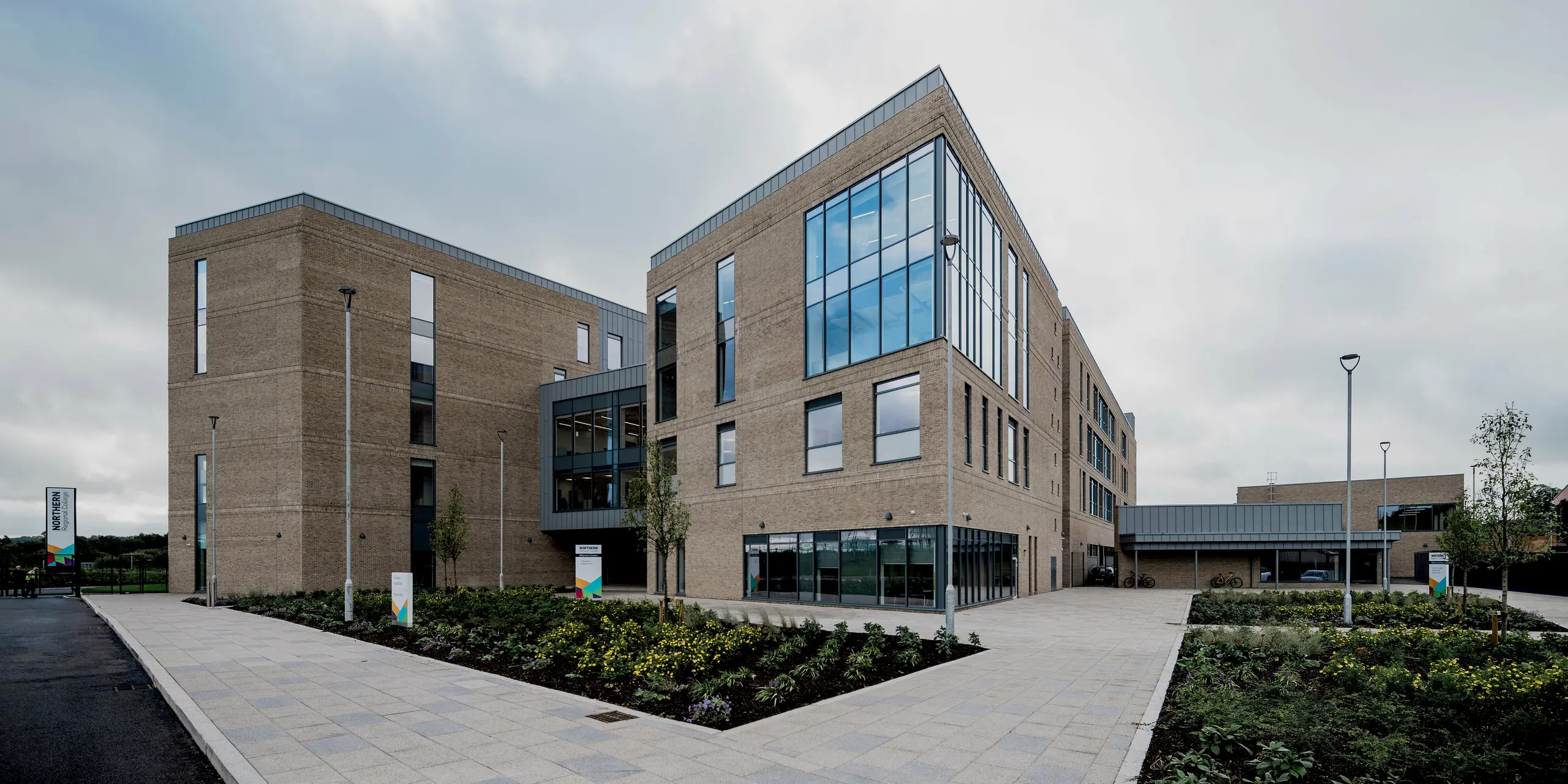 Une vue d'ensemble du Northern Regional College de Ballymena révèle plusieurs bâtiments à l'architecture bien définie, construits en briques claires et reliés par des surfaces vitrées et des passerelles ouvertes. Le long des toitures et des extrémités des façades, des bacs en aluminium PREFALZ à joints debout, couleur gris quartz, créent des accents horizontaux précis. Les surfaces métalliques encadrent élégamment l'architecture et contrastent avec la maçonnerie et l'immensité du ciel. L'aluminium PREFA est idéal pour les établissements scolaires grâce à sa durabilité, son faible besoin d'entretien et son design résolument moderne.