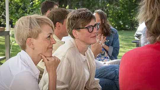 Gros plan sur Veronika Roll (Marketing PREFA) et Juliane Croce (photographe) lors de la table ronde d'experts PREFARENZEN 2027, organisée dans le jardin d'une résidence de vigneron. Assises, concentrées et attentives, les deux femmes participent à une discussion animée sur les projets architecturaux modernes. La scène, baignée de lumière naturelle, est agrémentée de verdure estivale et baignée d'une atmosphère détendue. À l'arrière-plan, on aperçoit les produits en aluminium PREFA, symboles de toitures et de façades durables et esthétiques, alliant fonctionnalité et design. L'image met en lumière l'échange créatif et l'intérêt profond du jury pour les idées de design, ainsi que l'inspiration suscitée par les solutions aluminium innovantes et de haute qualité de PREFA pour les projets d'architecture et de construction.