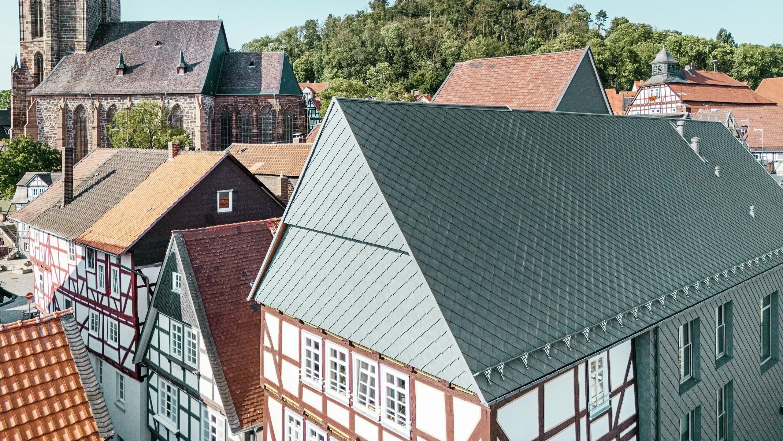 Le toit de la Volkshaus rénovée à Homberg, DE, avec des losanges 29x29 en aluminium PREFAen P.10 gris pierre, intégré dans la vieille ville historique avec des maisons à colombages, une église et un château. Le revêtement en aluminium durable et robuste allie un design moderne au charme de l'environnement historique. Au centre de l'image, on voit le pignon du bâtiment, à l'arrière-plan l'église et la colline du château.