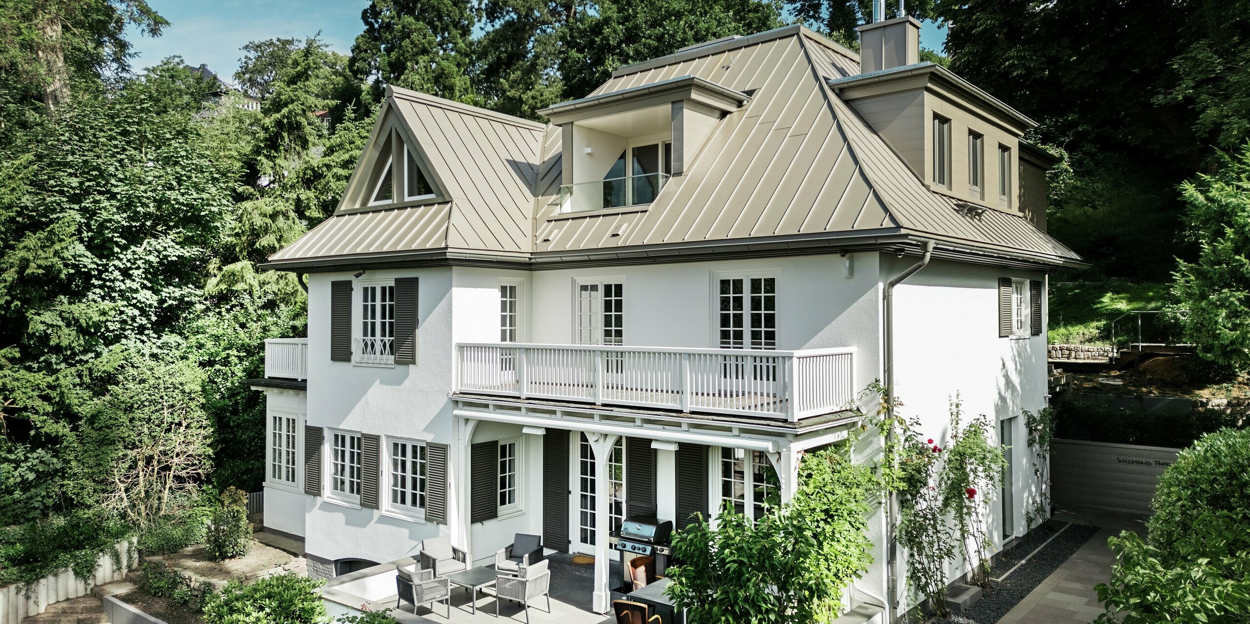 L'image présente la Villa Blankenese de face et légèrement en perspective oblique, nichée dans un écrin de verdure. La remarquable toiture PREFALZ couleur bronze souligne l'architecture et confère au bâtiment une élégance résolument moderne. La technique de la toiture à joints debout met en valeur la pureté des lignes et garantit une solution durable et résistante aux intempéries. Les bardages Sidings PREFA couleur bronze habillent les lucarnes et les éléments de façade, contribuant à l'harmonie de l'ensemble. L'alliance de produits en aluminium haut de gamme et d'une architecture classique fait de cette villa un projet d'exception.