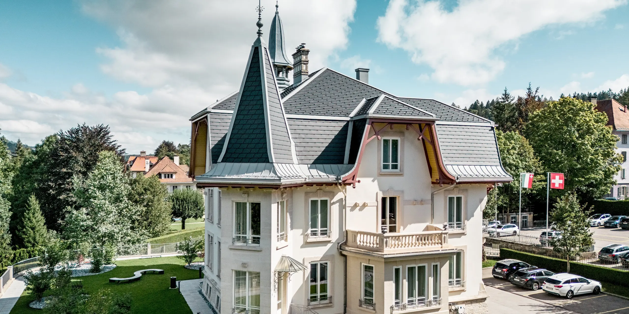 Spektakulärer Blick auf die Manufacture Horlogère du Locle in der Schweiz mit ikonischem Turm und kunstvoll modellierter Dachlandschaft. Die steil geneigten Flächen mit PREFA Dachschindeln in P.10 Schwarz erzeugen ein markantes, rhythmisches Muster, während PREFALZ in Patinagrau geschwungene Übergänge, Gauben und feine Anschlussdetails elegant formt. Das hochwertige Aluminiumdach überzeugt durch geringes Gewicht, hohe Witterungsbeständigkeit und außergewöhnliche Langlebigkeit – ideal für anspruchsvolle Architektur, die Design, Funktionalität und nachhaltigen Werterhalt perfekt vereint.