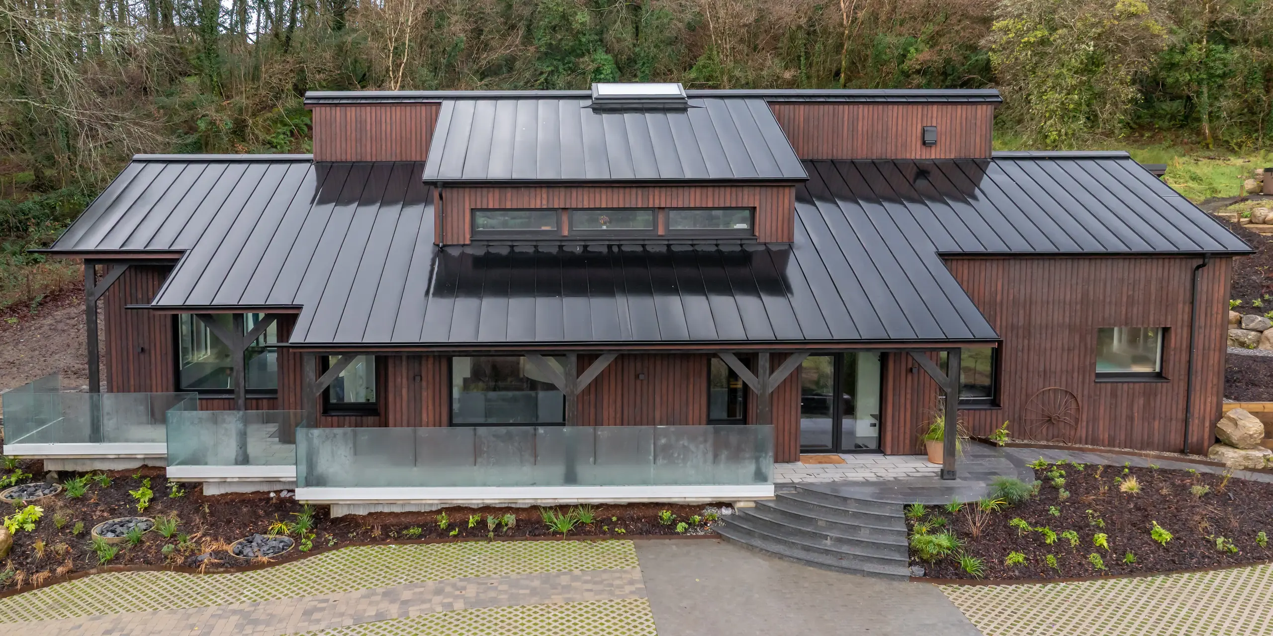 À Sligo, en Irlande, une maison individuelle moderne, nichée à flanc de colline, se distingue par sa façade en bois et son impressionnante toiture en aluminium PREFALZ P.10 Noir. La charpente apparente à joints debout se déploie avec dynamisme sur plusieurs pans de toiture, créant un effet architectural saisissant. De larges baies vitrées, des terrasses et des lignes épurées soulignent le design. La toiture en aluminium PREFA impressionne par sa durabilité, sa résistance à la corrosion et sa grande robustesse face aux intempéries.