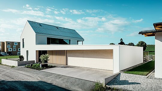 [Translate to Français:] Das Bild zeigt das moderne Einfamilienhaus in Freistadt, das sich durch seine schlichte, geometrische Form und die Kombination von reinweißer PREFALZ-Aluminium-Fassade und Holzvertäfelungen auszeichnet. Die Holzverkleidung am Garagentor bietet einen warmen, natürlichen Kontrast und fügt sich perfekt in das moderne Design ein. Das Dach, ebenfalls mit PREFALZ gedeckt, sorgt für eine nahtlose, wetterfeste Oberfläche. Diese Kombination aus pflegeleichtem Aluminium und Holz schafft eine ästhetisch ansprechende und funktionale Lösung für nachhaltige Architektur. Der weitläufige, grüne Außenbereich und der klare Himmel im Hintergrund unterstreichen die moderne Lebensqualität, die dieses Haus bietet.