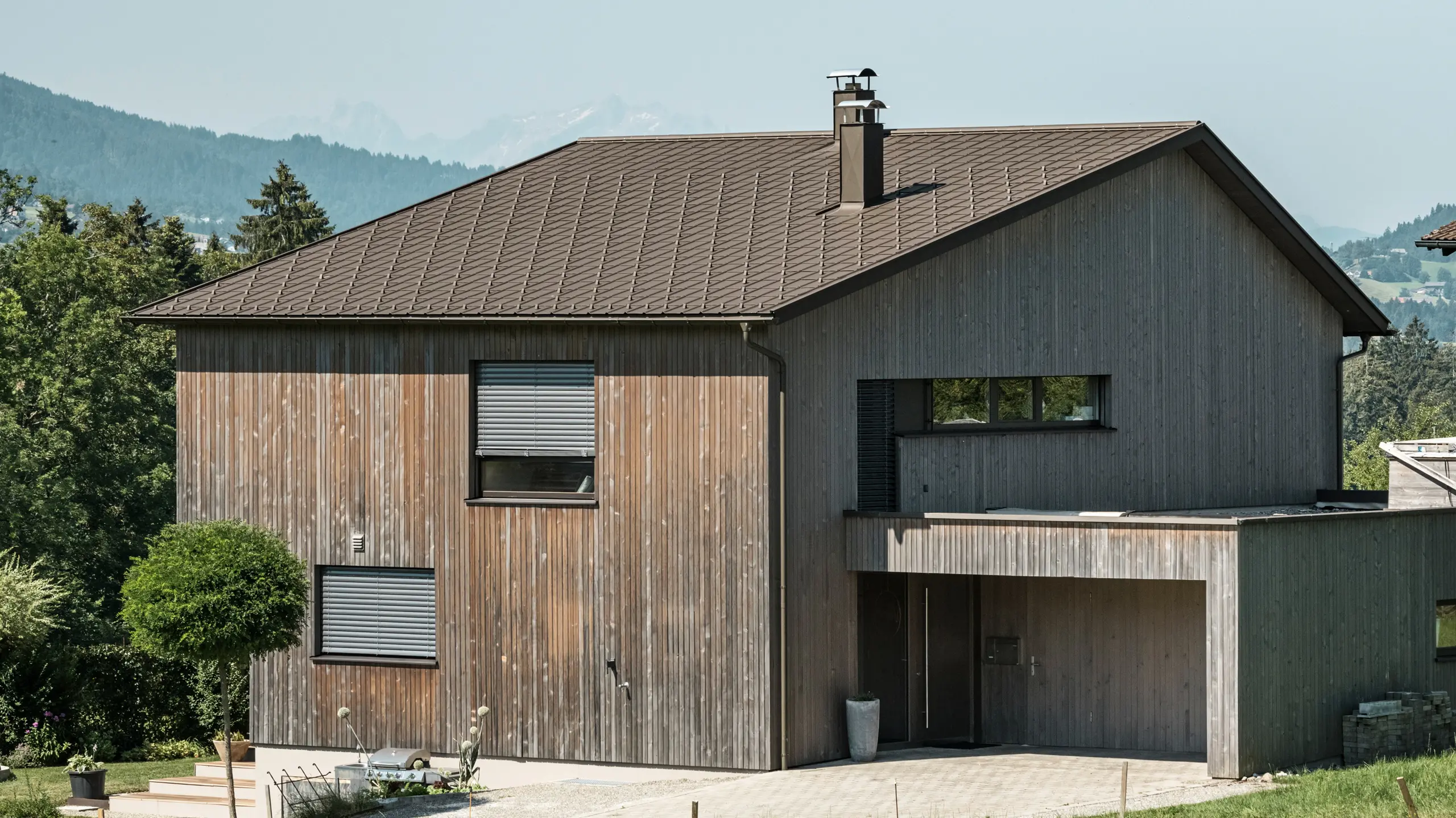 Vue latérale d'une maison individuelle contemporaine à Krumbach, avec une façade en bois naturel et une toiture en aluminium PREFA durable de couleur P.10 brun. L'harmonie des couleurs montre l'intégration élégante d'un design moderne et de matériaux traditionnels, tandis que les tuiles PREFA assurent une conception esthétique et fonctionnelle de la toiture.
